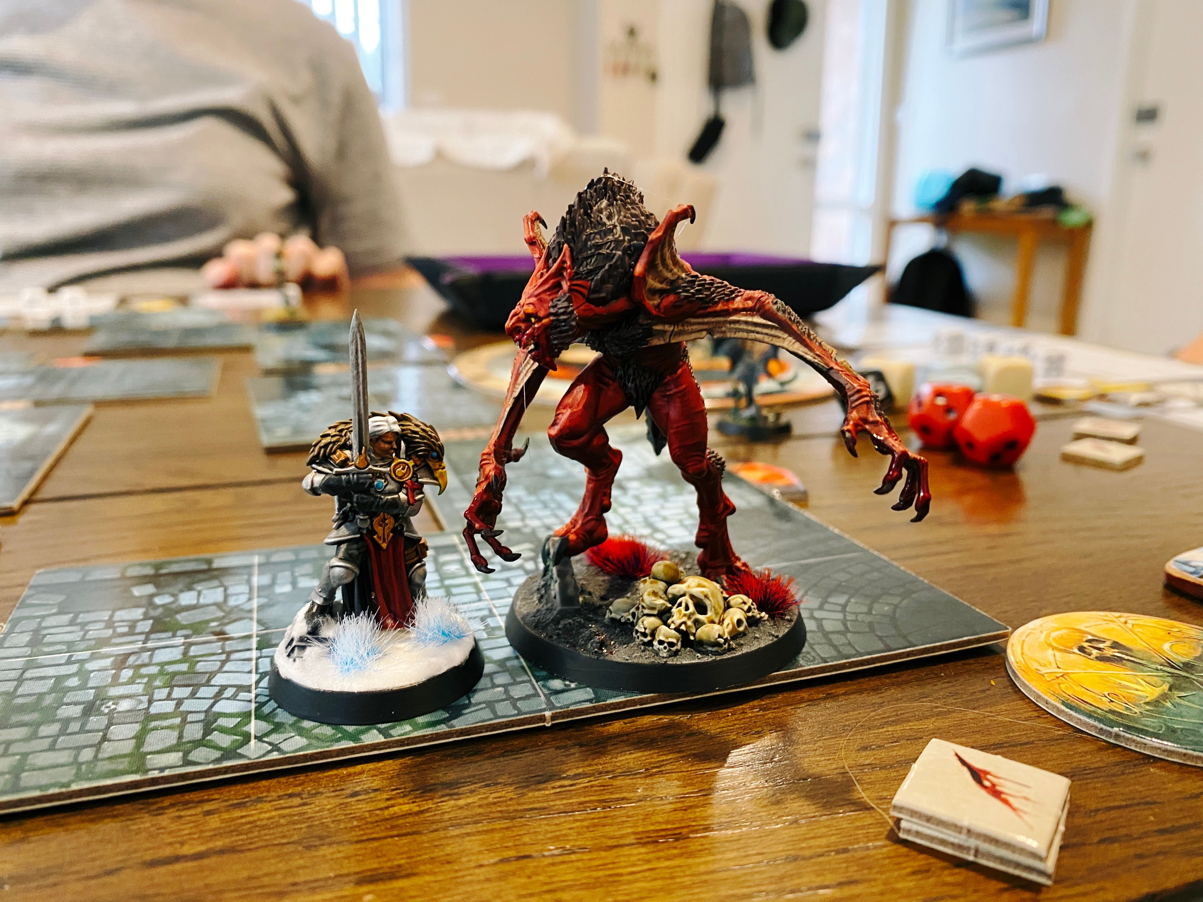 A photo of two painted miniatures standing next to each other, one is a heavily armoured warrior woman with short hair holding a large sword, and positively LOOMING over her is the Vargskyr, which is a massive hunched over demon/bat hybrid thing twice the height of the warrior, with red skin and brown fur down its arms. It has blood around its mouth and dripping from its claws and there's a pile of skulls on its base.
