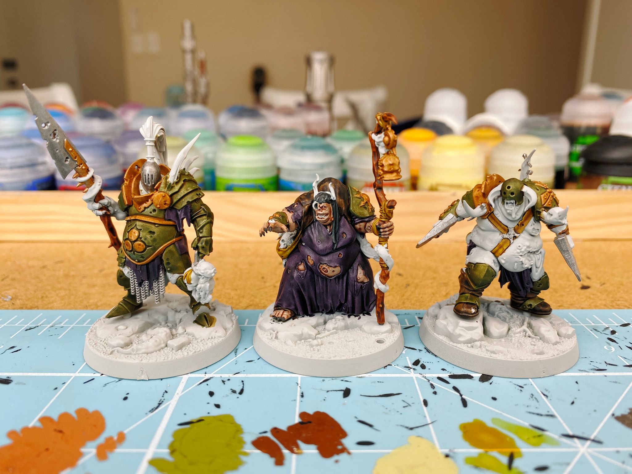 A photo of 3 partially-painted Warhammer Underworlds miniatures. One is heavily armoured and has a spear in one hand and a rack of severed heads in the other, one is corpulent and in ragged robes with holes in them, holding a staff in one hand and has her other hand out like she's casting a spell, and the 3rd is also corpulent with a gash on the side of his stomach and his insides spilling out. He's roaring and holding two axes.