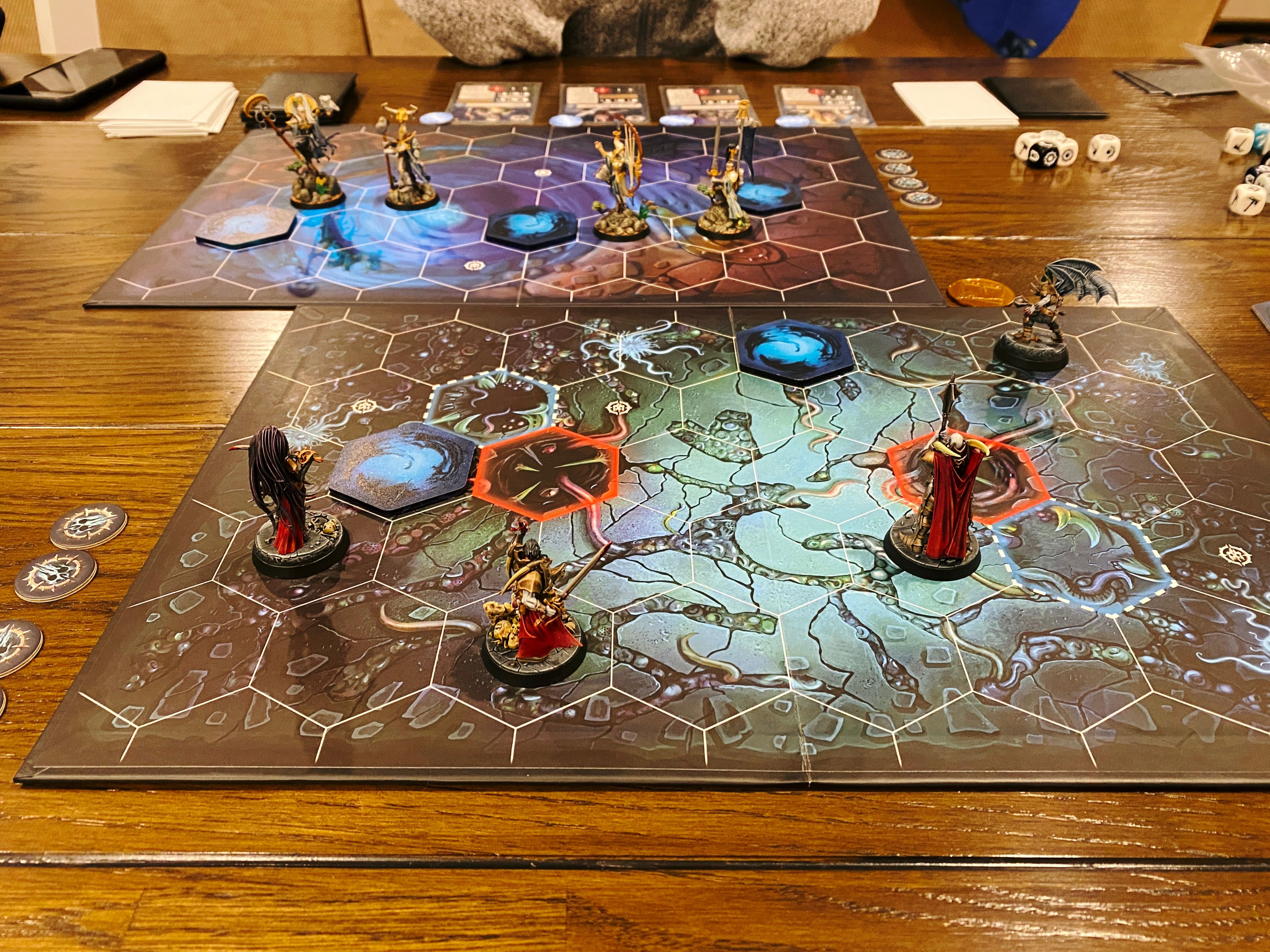 A photo of the initial setup of a game of Warhammer Underworlds. It's a miniatures board game played on boards with hexagonal tiles, closest to the camera are four armoured vampires with pale blue skin and red cloaks, with blood dripping from their weapons and the corners of their mouths, and on the other side are four lithe and lightly-armoured elves.