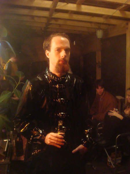 A photo of me, a white man with short hair and a long red goatee, wearing an EXTREMELY goth coat PVC-and-buckles coat.