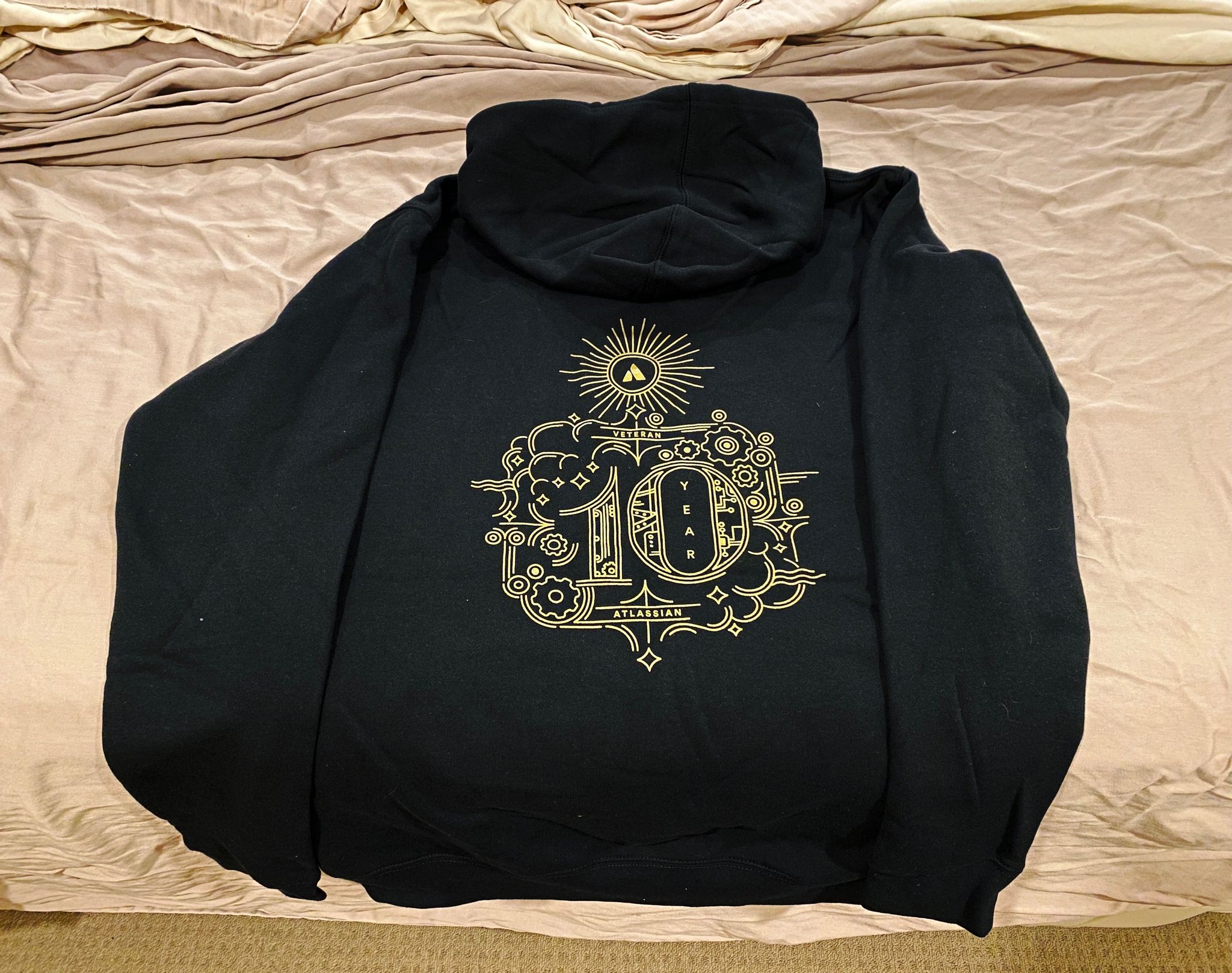 A photo of the back of a black hoodie sitting on a bed. It says "10 year" on it with clouds and cogs around it, all in a stylised outline line design in a gold colour.