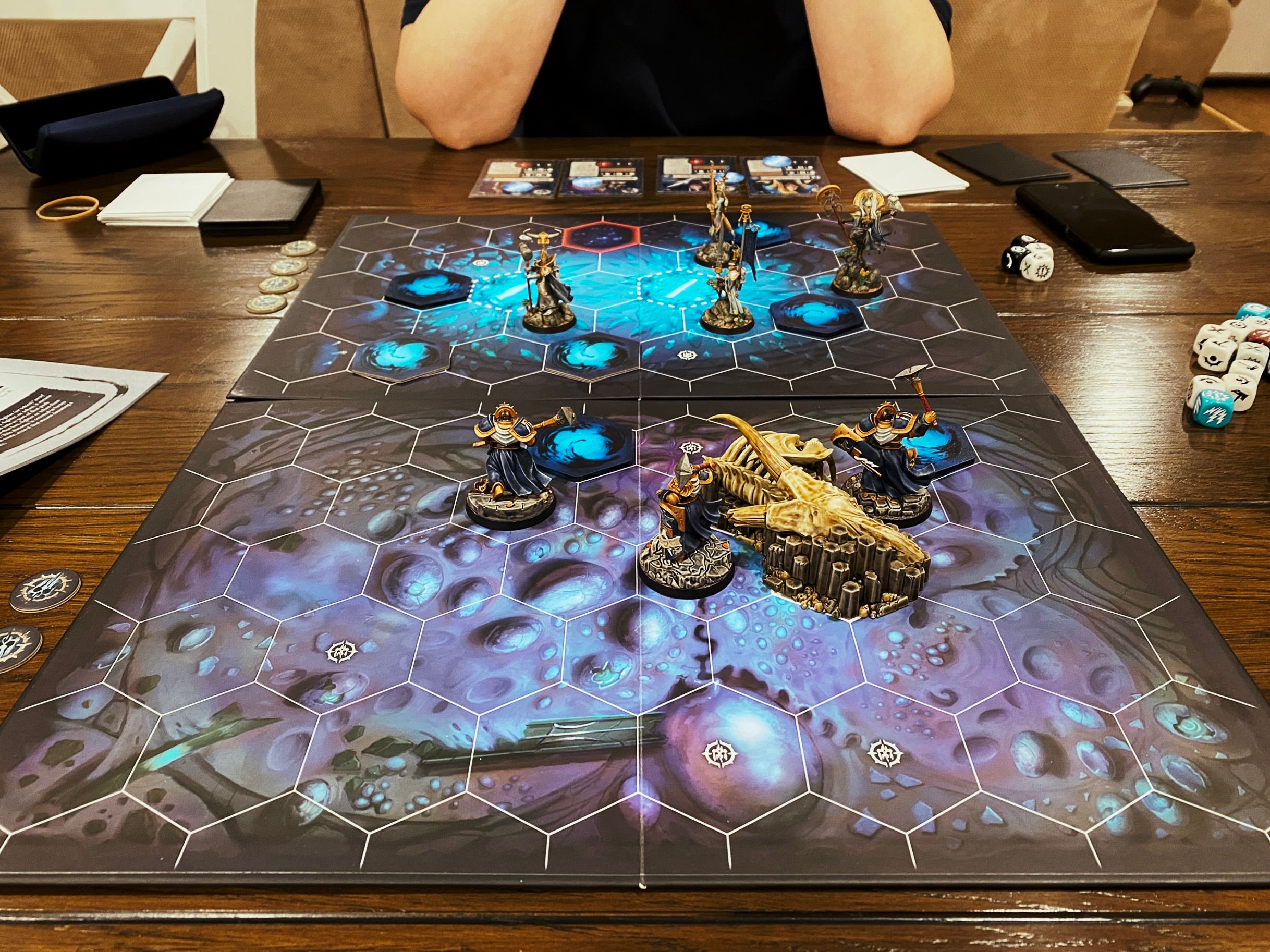 A photo of the board of a Warhammer Underworlds game. The board has hexagonal tiles printed on it with a blue murky-depths kind of feel of artwork, at the front are three heavily-armoured warriors in gold and dark blue armour (Ironsoul's Condemnors), and at the back are four lithe and elegant aelves (Myari's Purifiers).