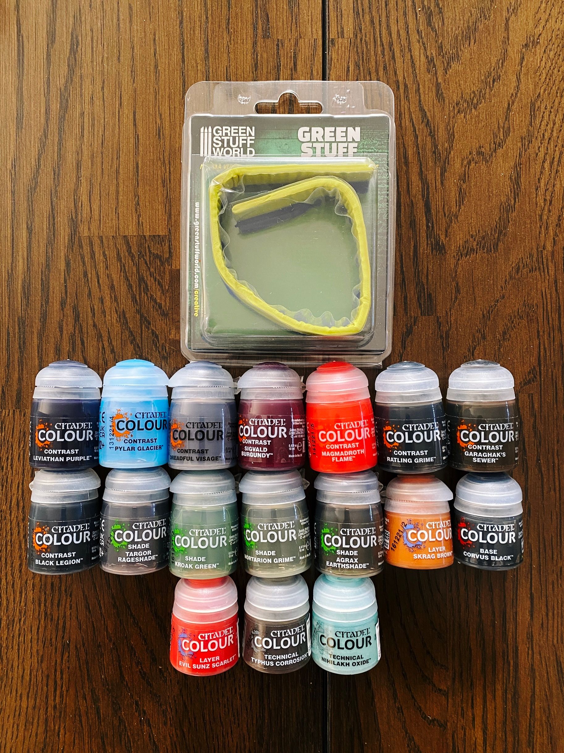 A photo of seventeen little pots of miniature paint, plus a roll of "Green Stuff" which is used for modelling and filling gaps in miniatures.