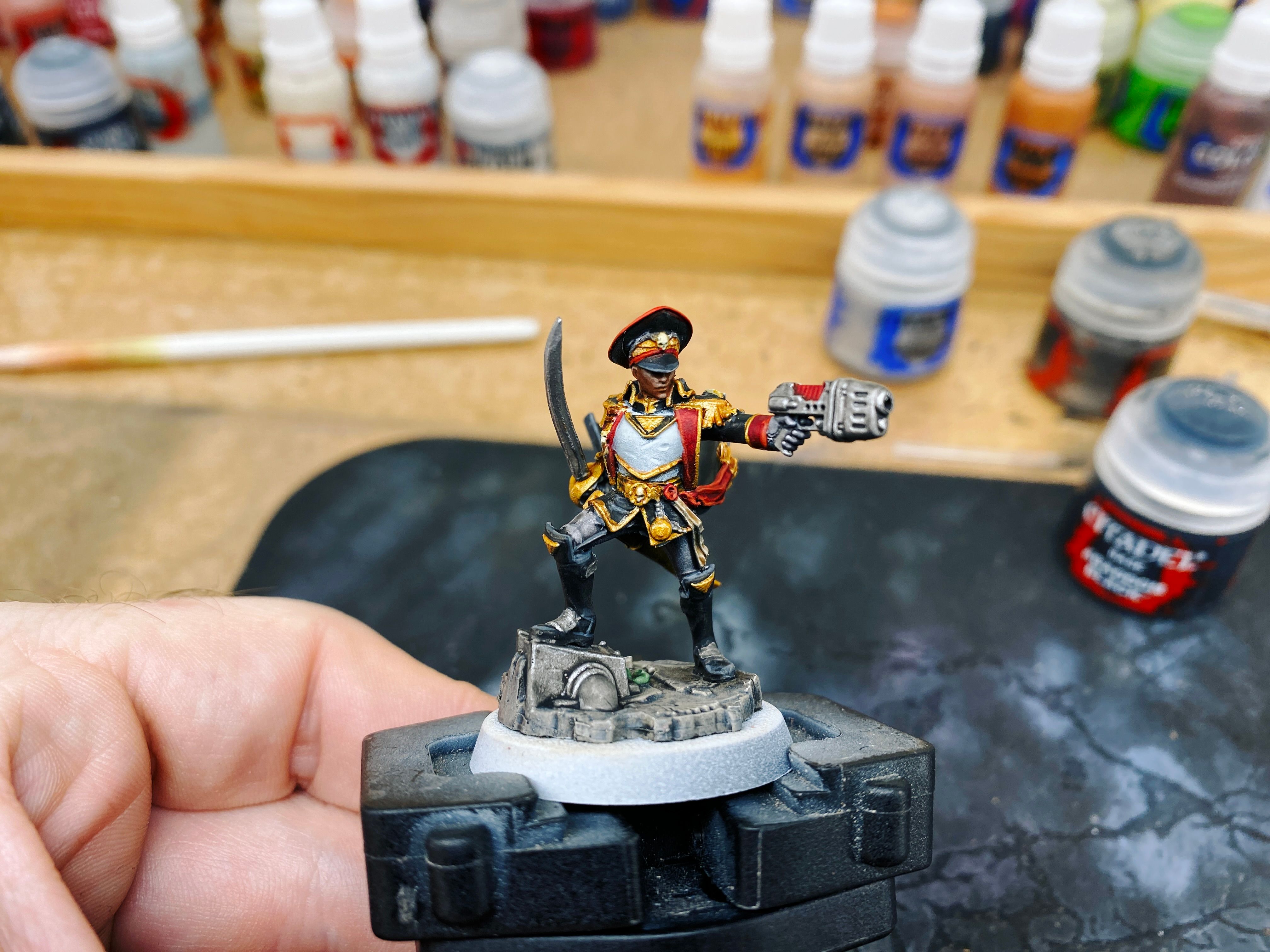 A photo of a mostly-painted Warhammer 40,000 miniature wielding a plasma pistol and sword and standing in a very heroic pose. She's got a military cap on and her black stormcloak is billowing out behind her.