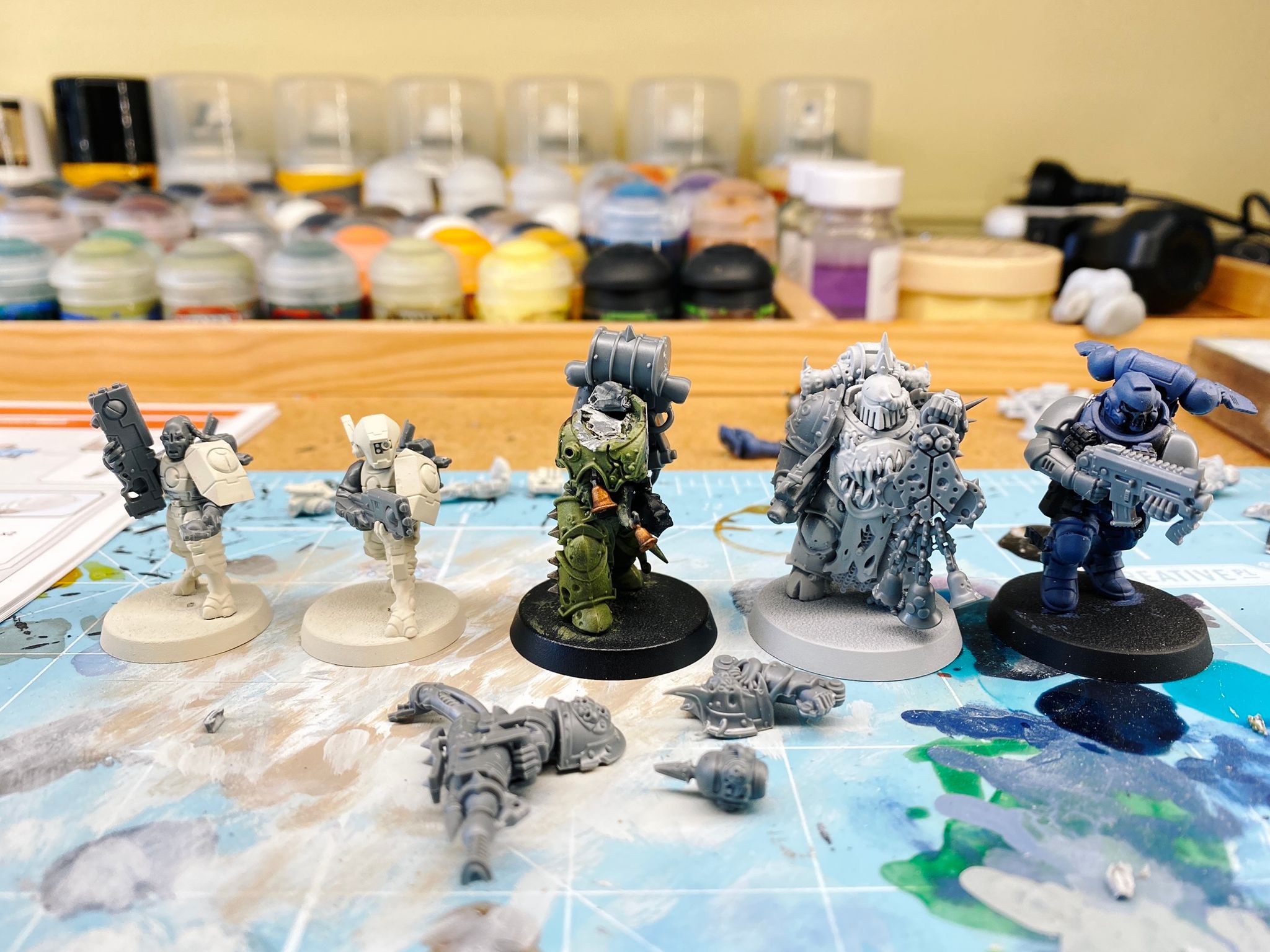 A photo of five miniatures (2 Tau, 2 Death Guard, 1 Space Marine Reiver) with an undercoat colour of white or black, with their weapons clearly newly-added because they're fully unpainted raw grey plastic. The miniature in the middle isn't fully assembled and is missing his head and both arms.