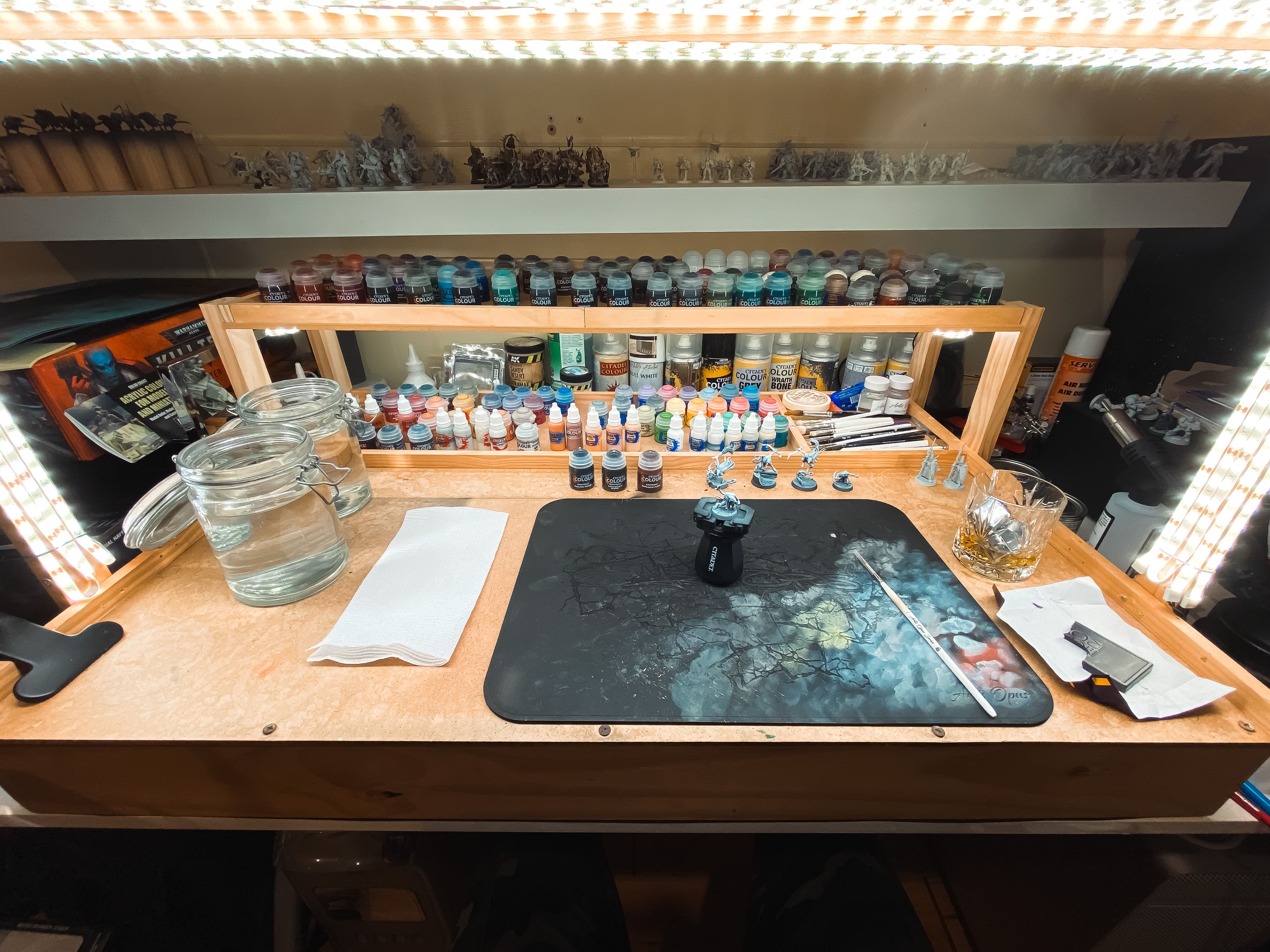 A photo of my painting desk setup, with an arch of LED strip lights illuminating the working area, five Skaven (rat-men) miniatures ready to be painted, and a bunch of little pots of paint spread over two shelves.