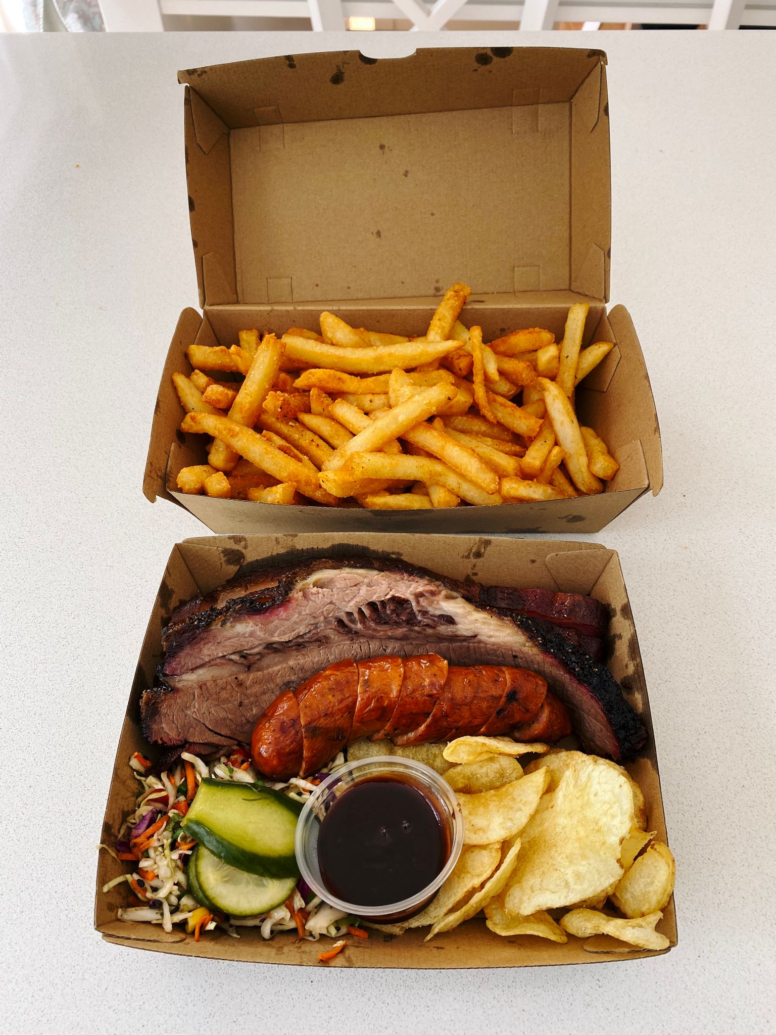 A photo of two takeaway food boxes, one with hot chips, the other with barbecued beef brisket, pork belly, sausage, chips, and slaw.