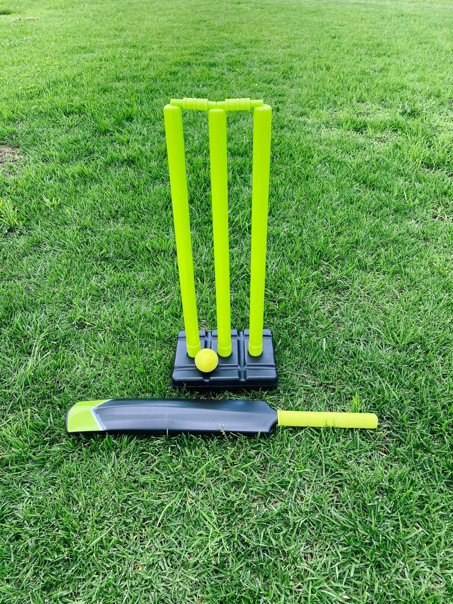 A photo of a fluro green set of plastic wickets, a ball, and a cricket bat sitting in a grassy field.