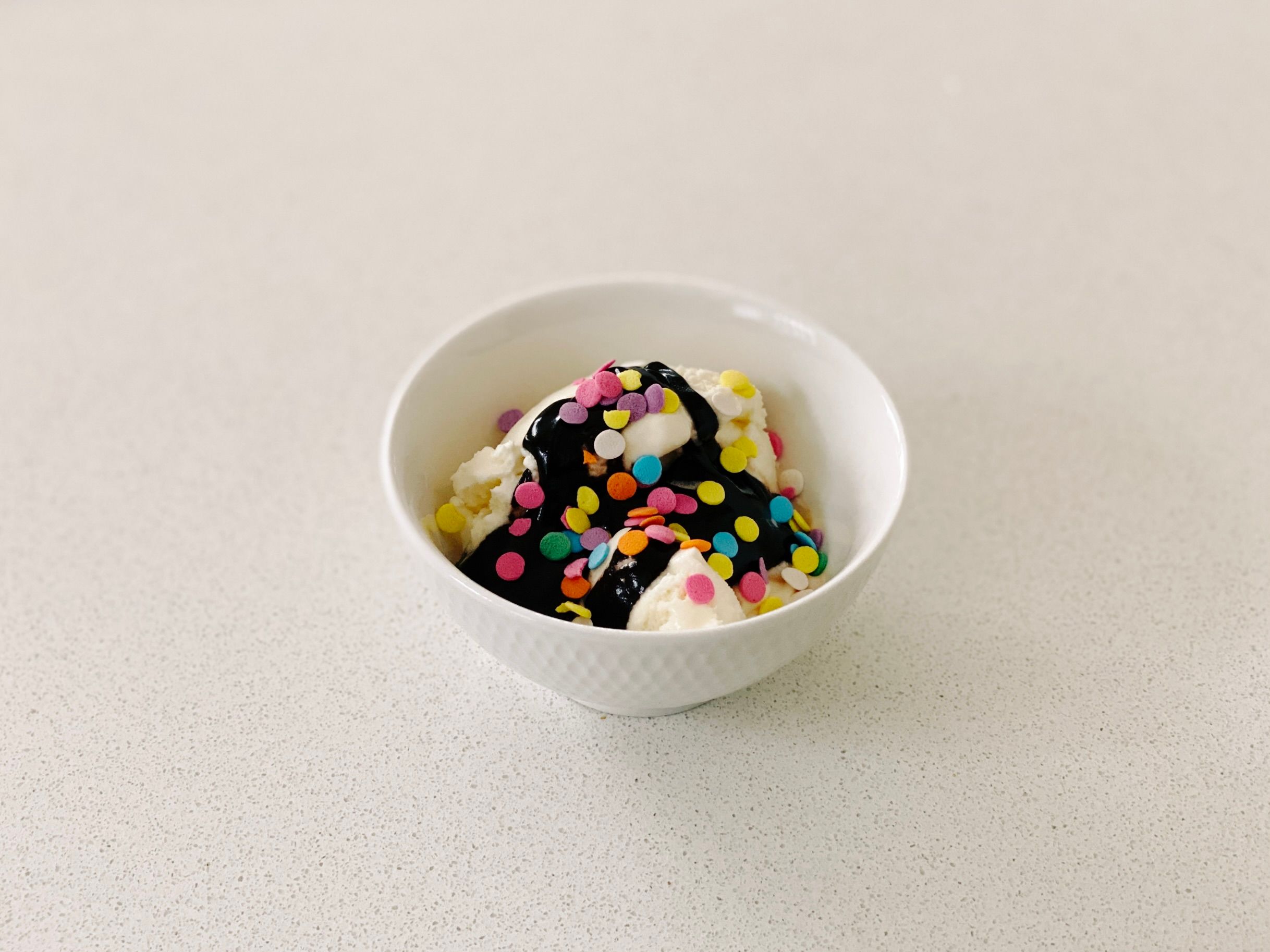 A photo of a little bowl of ice cream with hot fudge on top and multi-coloured sprinkles.