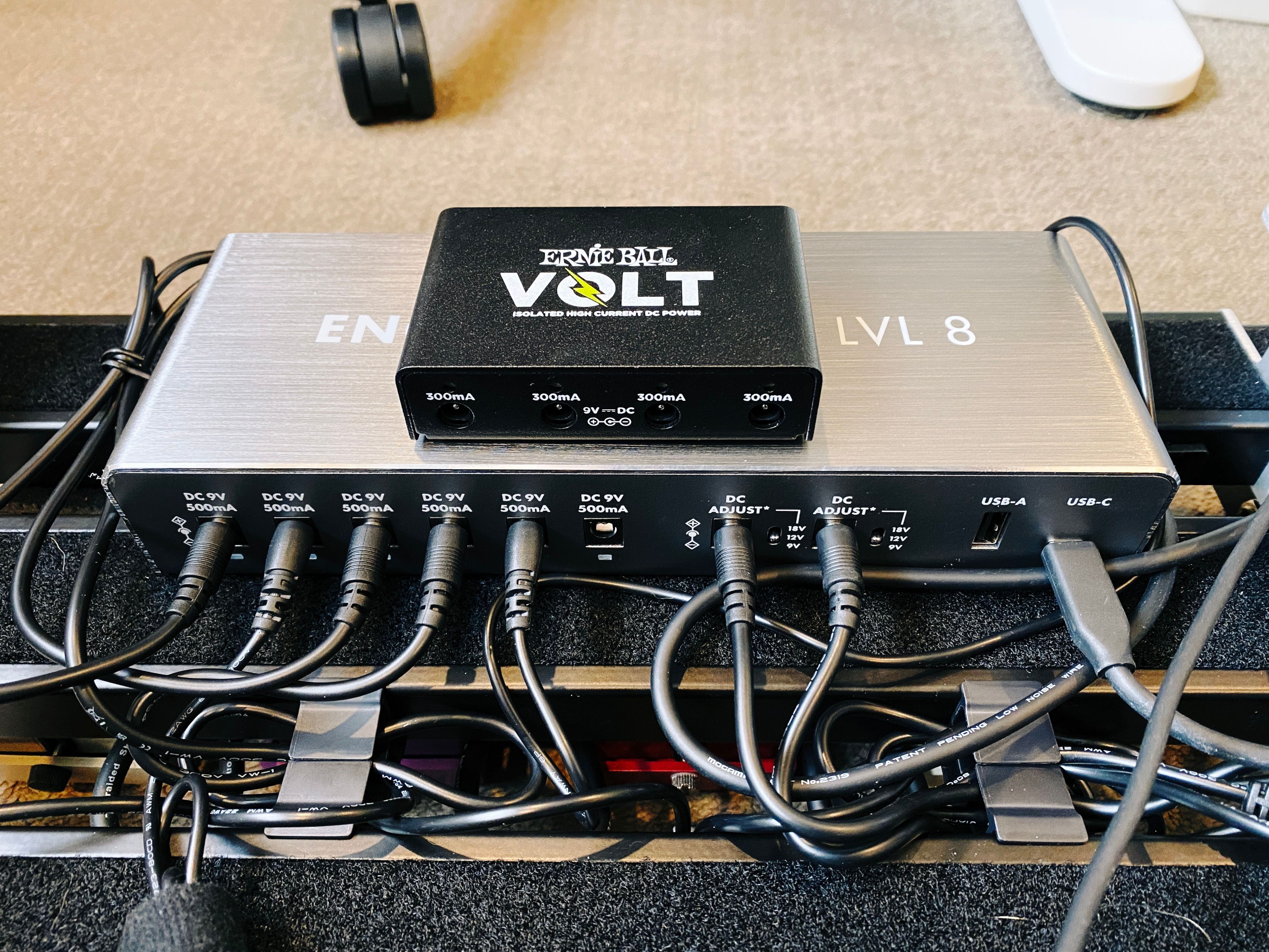 A photo of the bottom of my guitar pedal board. There's a large silver power supply attached to the bottom with lots of cables coming out of it, with a FAR smaller black one (the old one) sitting on top. Just eyeballing it, by volume I reckon the new power supply is probably like six or seven times larger, if not more.