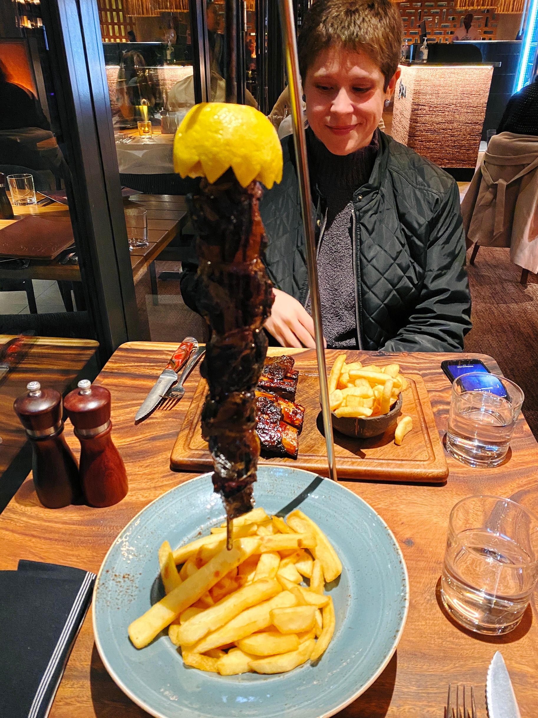 A photo of a metal contraption that has a skewer through multiple pieces of delicious-looking lamb and is hanging the skewer vertically above a plate of chips.