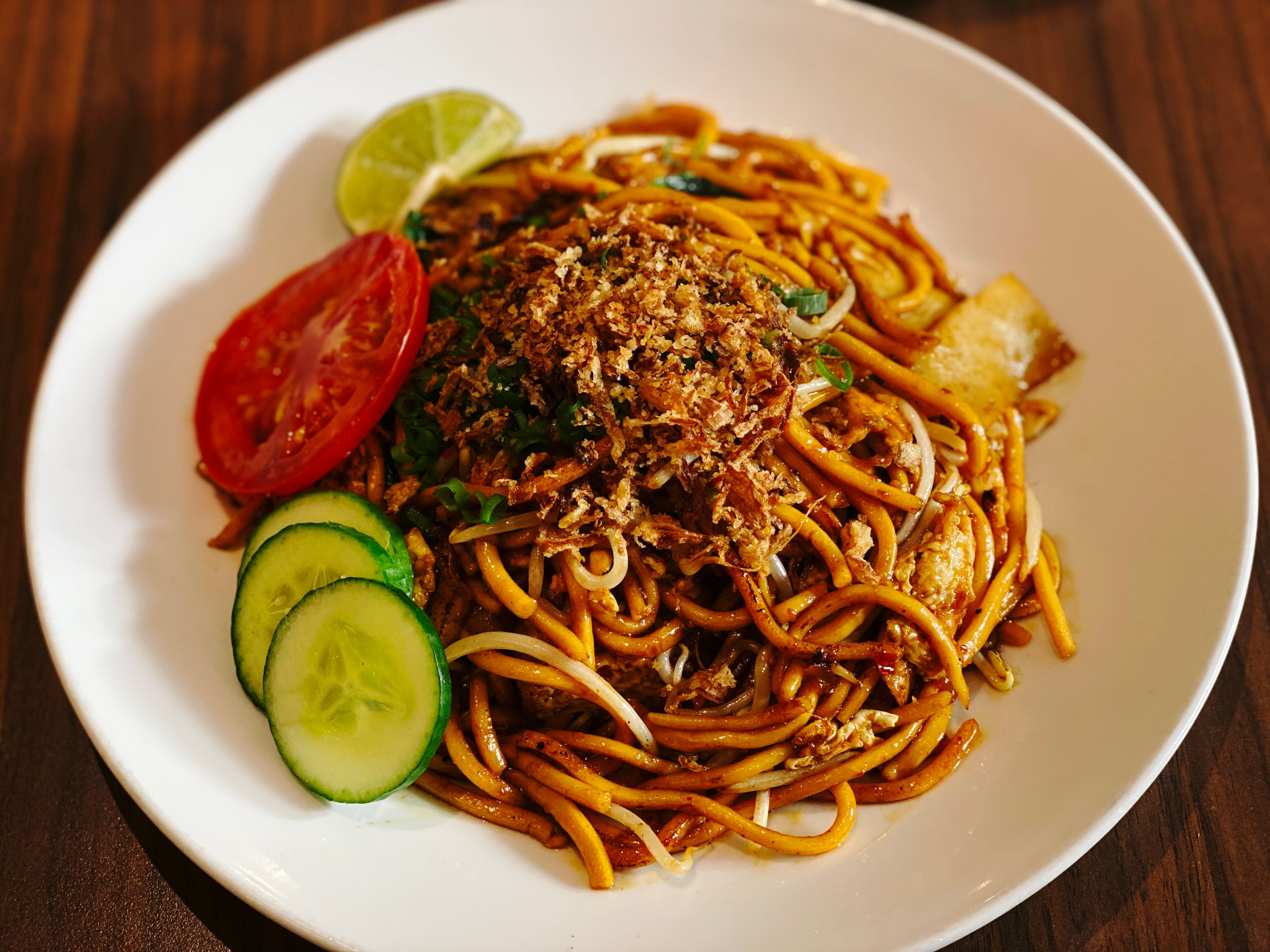 A photo of a plate of mee goreng, with a slice of tomato, lime, and three slices of cucumber next to it.