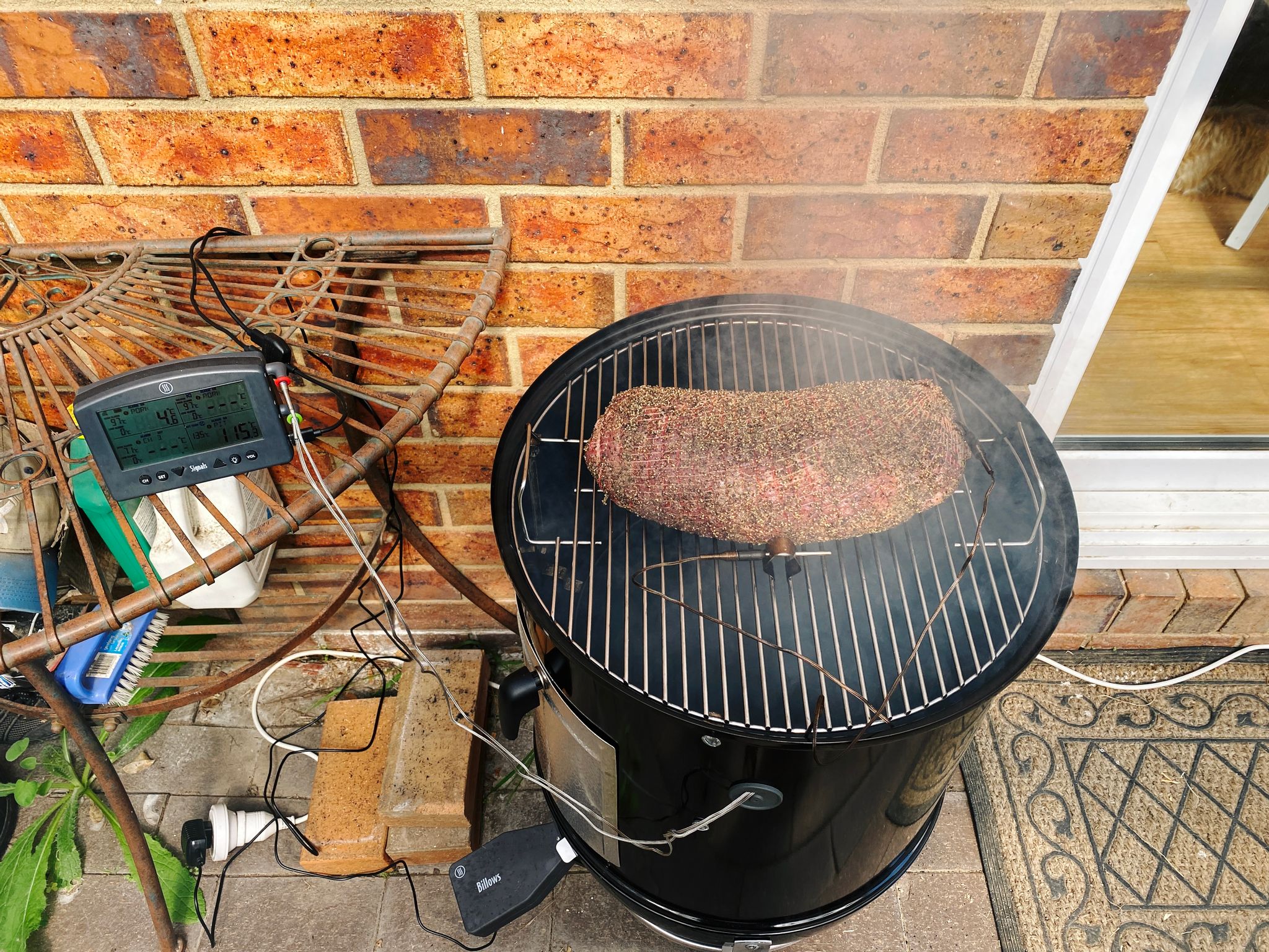 A photo of a pork collar butt sitting on a Weber Smokey Mountain smoker with smoke coming up from underneath. It has a temperature probe in it, and another to measure the ambient temperature inside the smoker.