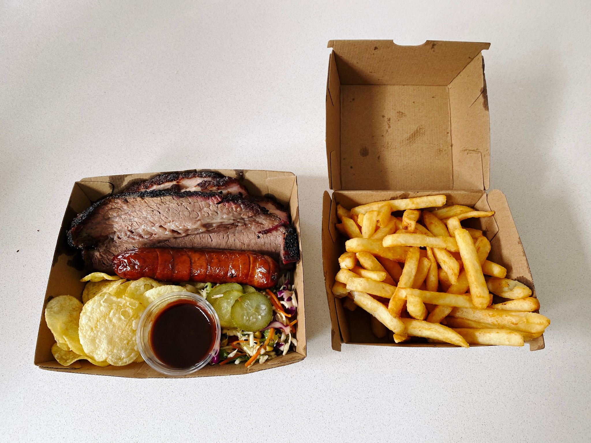 A photo of a takeaway box with beef brisket, pork belly, and sausage in it, as well as potato chips and some salady thing I didn't eat because it has coriander in it, and other box with hot chips in it. I am extremely full.
