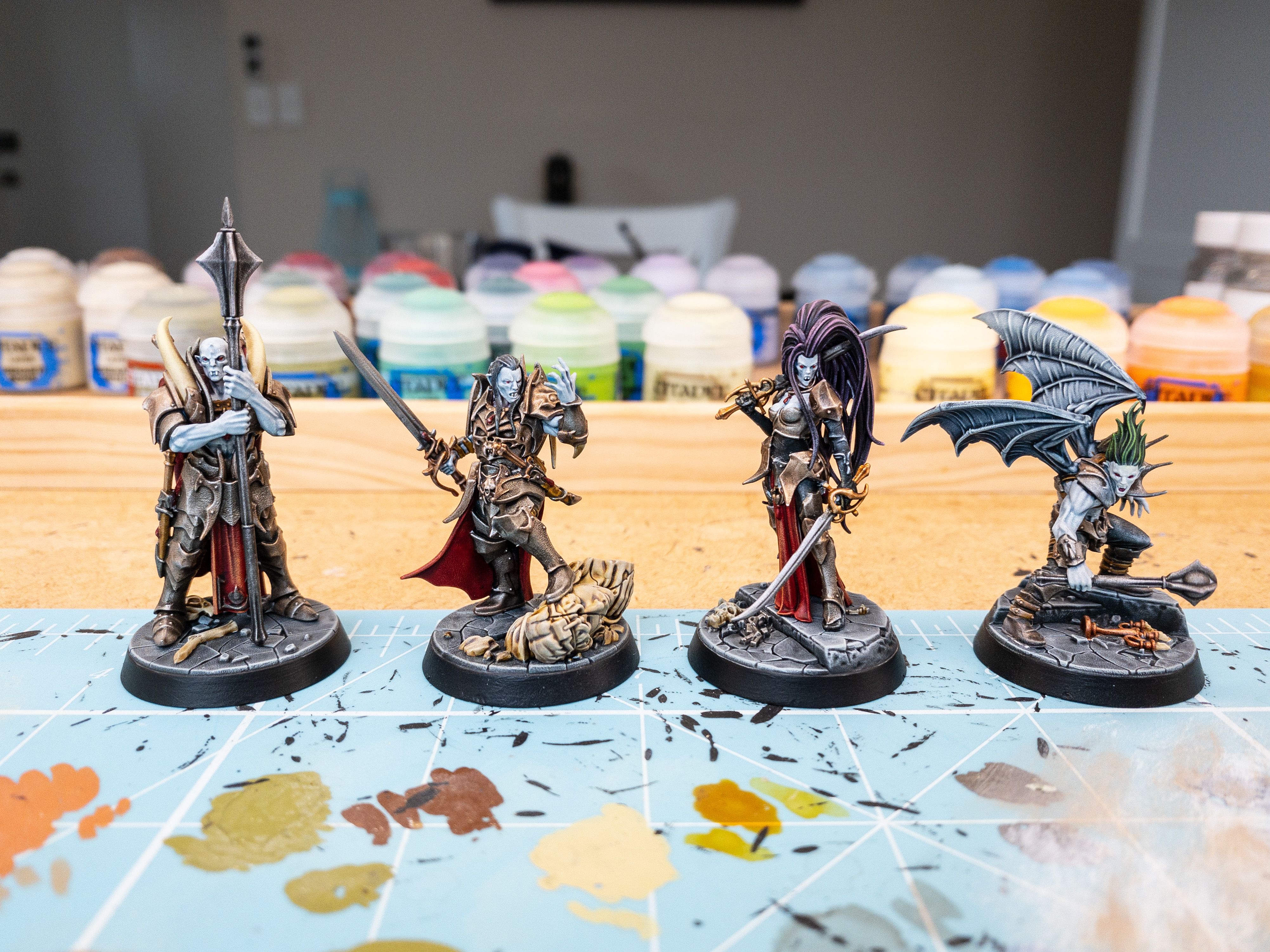A photo of four vampire miniatures, all heavily armoured. One is bald and is holding a huge mace vertically in front of him, the second has long black hair and he's wielding a sword and has one hand up in front of him as if he's doing a Darth Vader-style Force crush. The third is a very buxom lady in an armoured corset wielding two swords, with CRAZY long purple hair. The last is hunched over and looks like he's hissing, he's got a one-handed mace and big bat wings.