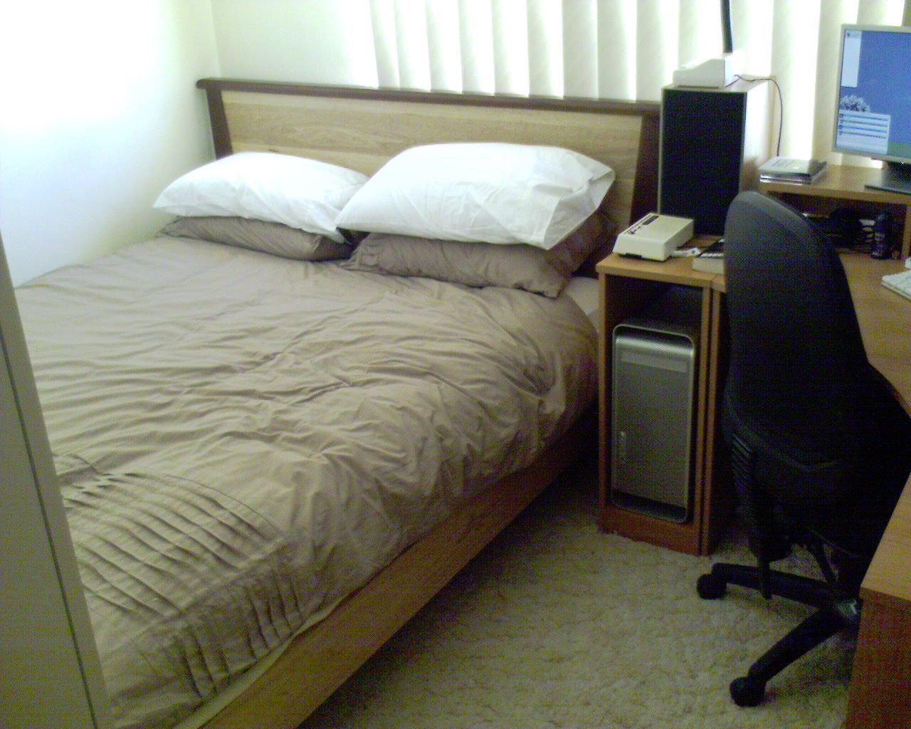 A photo taken from the doorway of a rather small room. There's a queen-size bed sitting on a wooden slatted frame, all neatly made up with pillows and blankets, alongside part of a corner desk with a computer on it.