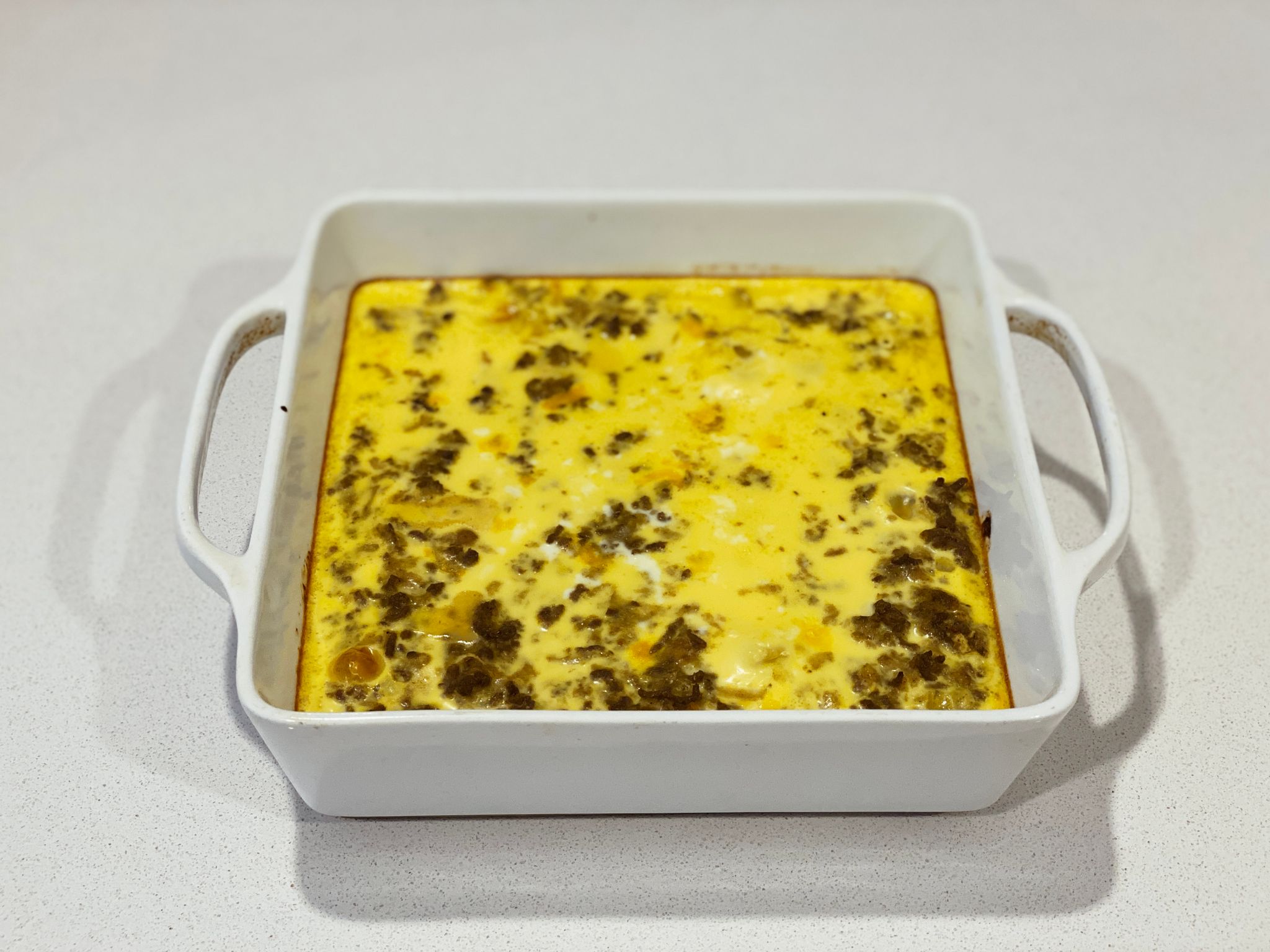 A photo of a white square ceramic oven dish with the finished bobotie. The top is very yellow where the egg is, and there's bits of beef mince poking through.