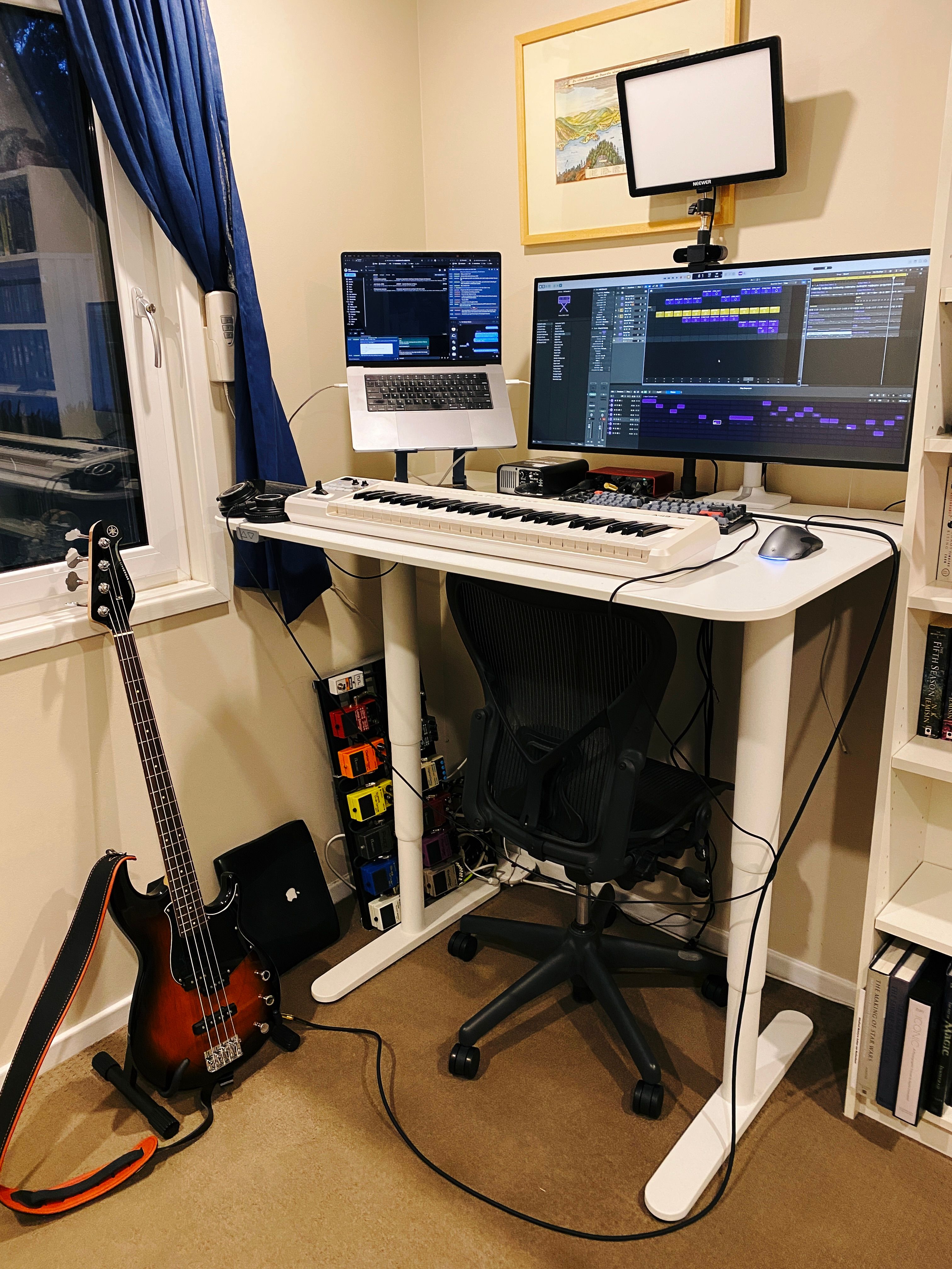 A photo of my standing desk in its upright position, with a bass guitar sitting to the left of the desk, a MIDI keyboard on the desk, and Logic Pro showing on the main display.