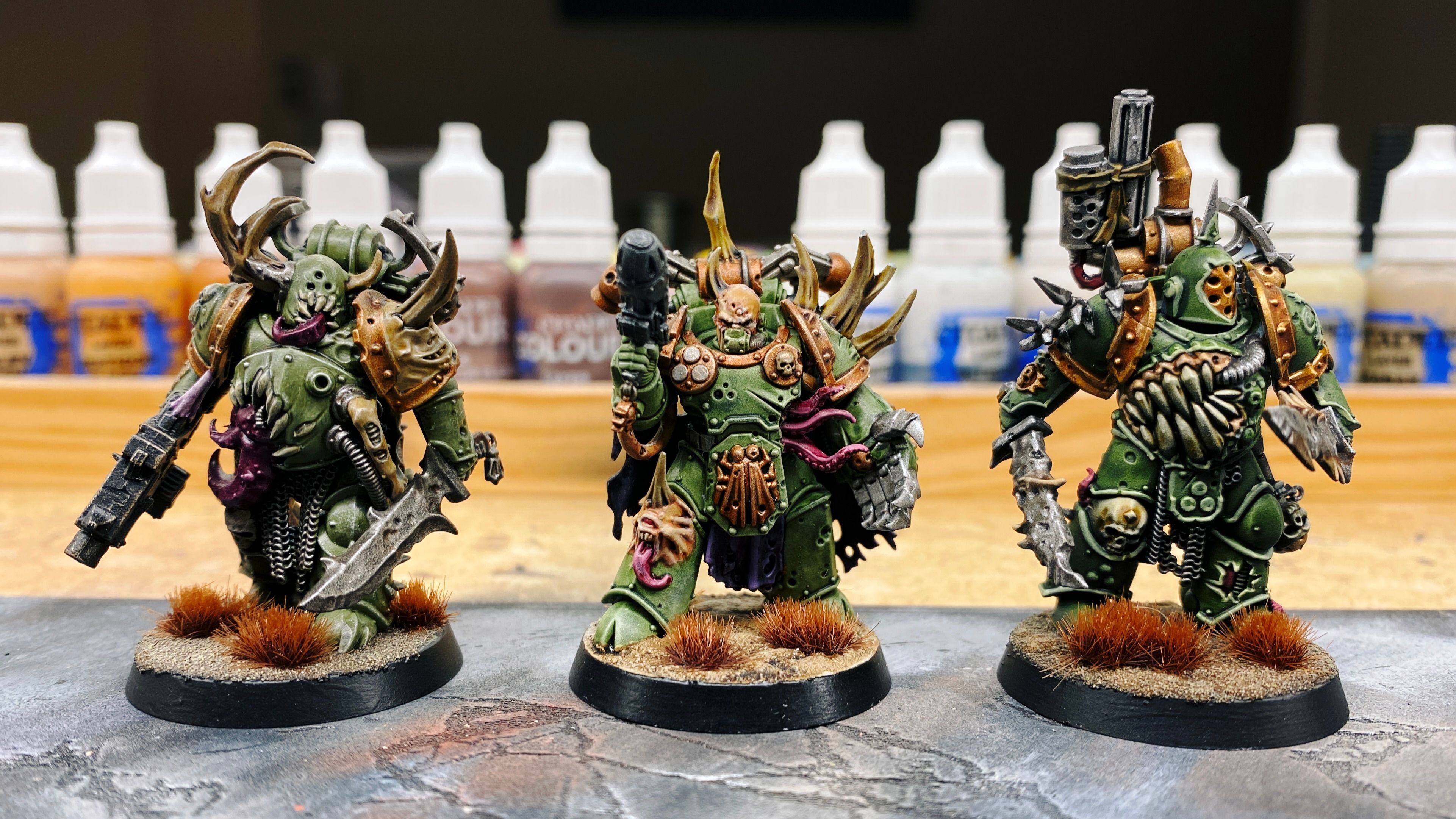 A photo of three Plague Marines in a line. They're all in the same general khaki green armour, but the one on the left is fairly dull-looking without too much highlighting since it was my old regular-paints painting technique where the highlighting was done with drybrushing. The one in the middle has come out a brighter green because the underlying primed colour is brighter and also it's the newer "Militarum Green" Contrast paint so the primed colour has a lot more effect on the finished product. And finally we have the one in the previous photo with the two knives that has much more emphasised shadows and much punchier highlights thanks to the zenithal highlight and manual highlighting.