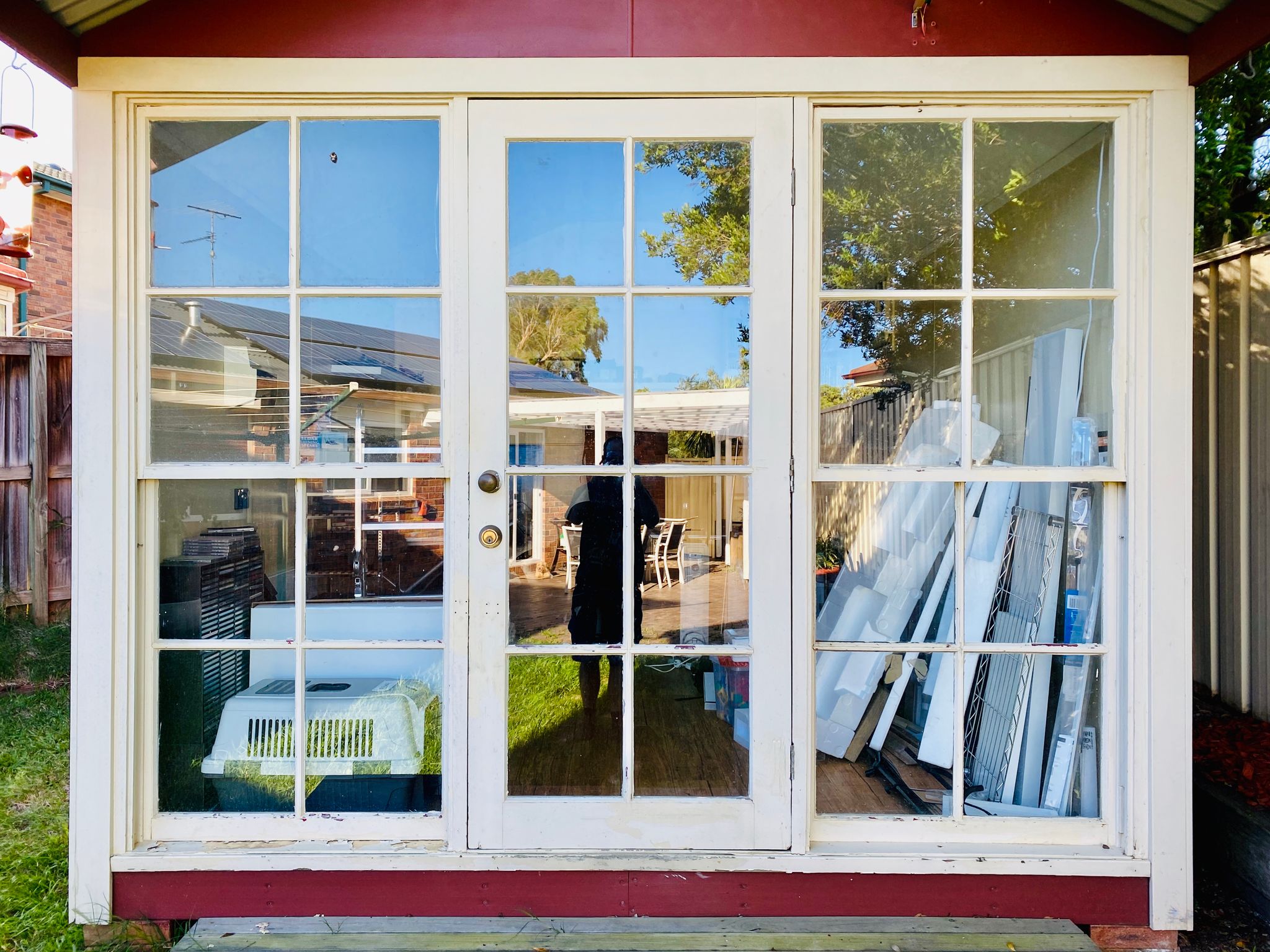 A photo of the front of a stand-alone backyard room. The front of it is primarily glass with a painted wood frame (door in the middle, two full-length windows on either side), and the paint is peeling and the wood isn't in very good condition.