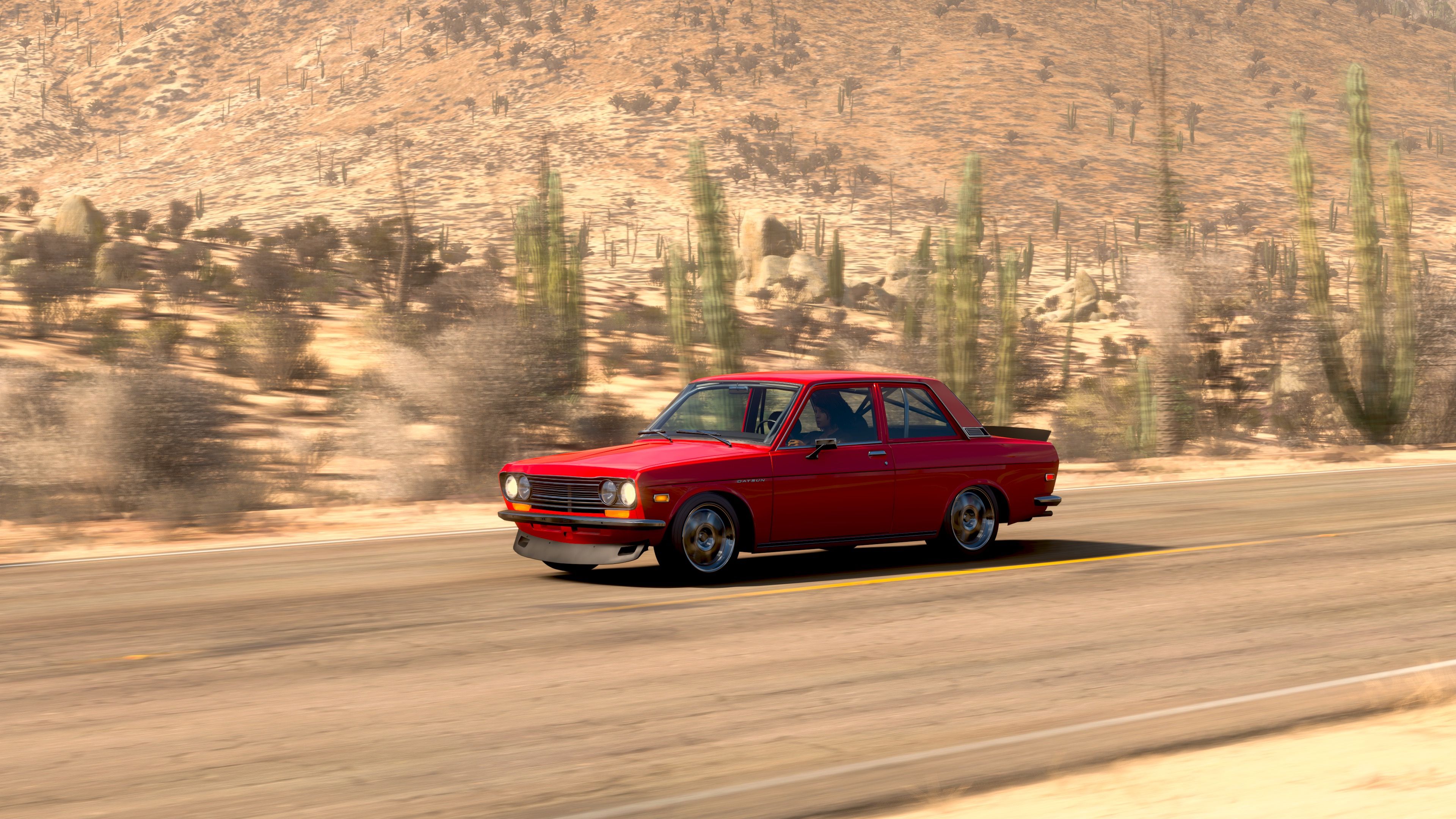 Another screenshot from Forza Horizon 5, showing my red Datsun 510 with big rims zooming along the road in the middle of the desert, there's lots of cactii and scrubby bushes on the side of the road.