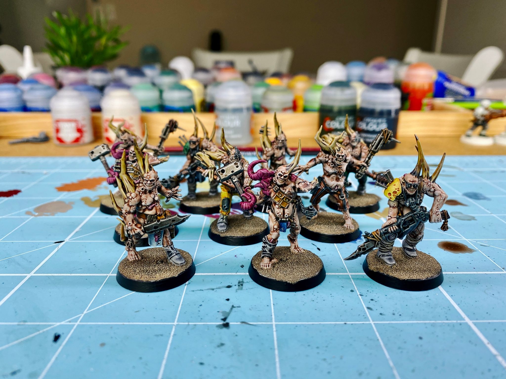 A photo of ten fully-painted Nurgle poxwalkers. They're gross distended zombie-looking things with big bone spikes growing out of them, some have pink tentacles, there's lots of pustules all over their bodies, and they're brandishing all sorts of makeshift pieces of industrial equipment as weapons.
