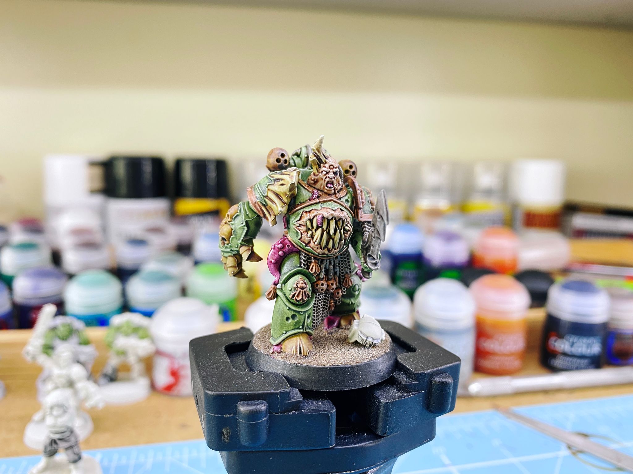 A photo of a Plague Marine from Warhammer 40,000. He's got green armour with skulls in various places, plus horns ripping through the armour on one shoulder and purple tentacles coming out from various armour joints. His head is corpulent and has pustules on it, and his abdomen has a large mouth with teeth bursting through the armour there. On the base is a white Imperial Guard helmet lying on the ground as though he's just killed them.