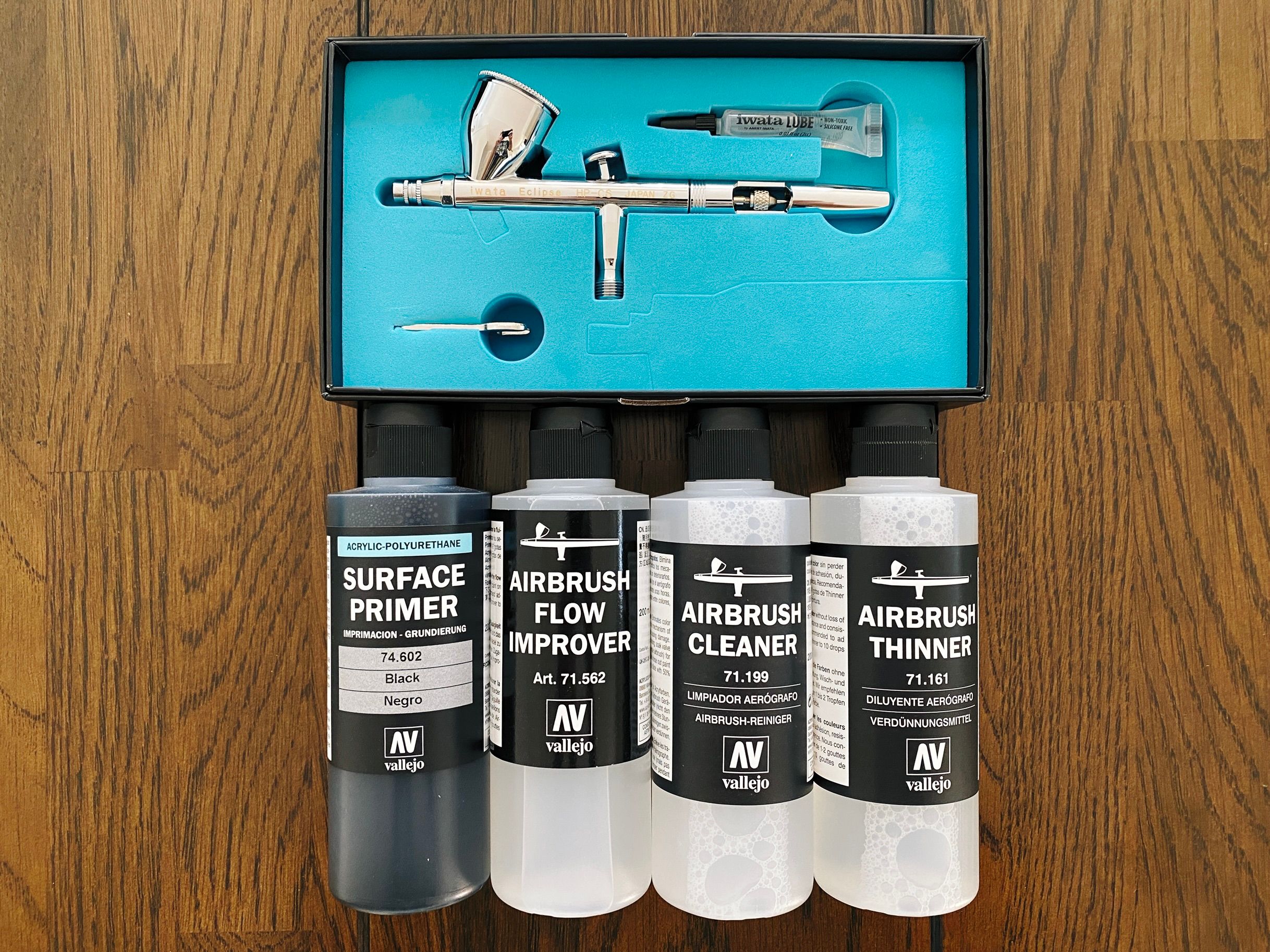 A photo of an Iwata Eclipse HP-CS airbrush sitting in its box, along with bottles Vallejo-brand black surface primer, airbrush thinner, airbrush flow improver, and airbrush cleaner.