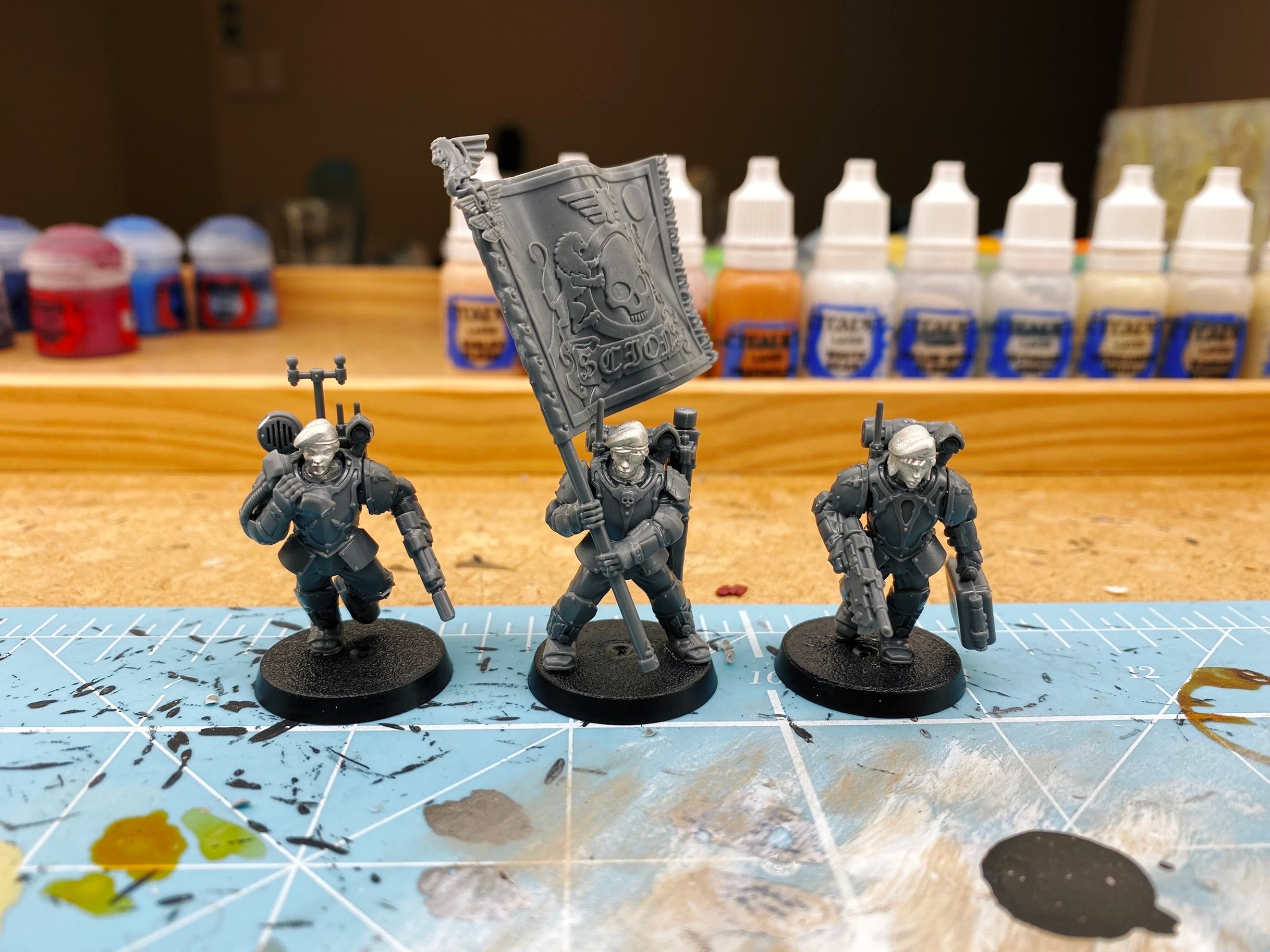 A photo of three miniatures in plain grey plastic, all heavily armoured. One has a chunky telephone handset and is yelling into it (she's the comms woman), one is holding a giant flag that is the regimental standard, and the third has a medkit bag in one hand and a pistol in the other. The heads of all three are shiny pewter instead of being plastic and are wearing berets, and they're all clearly female.