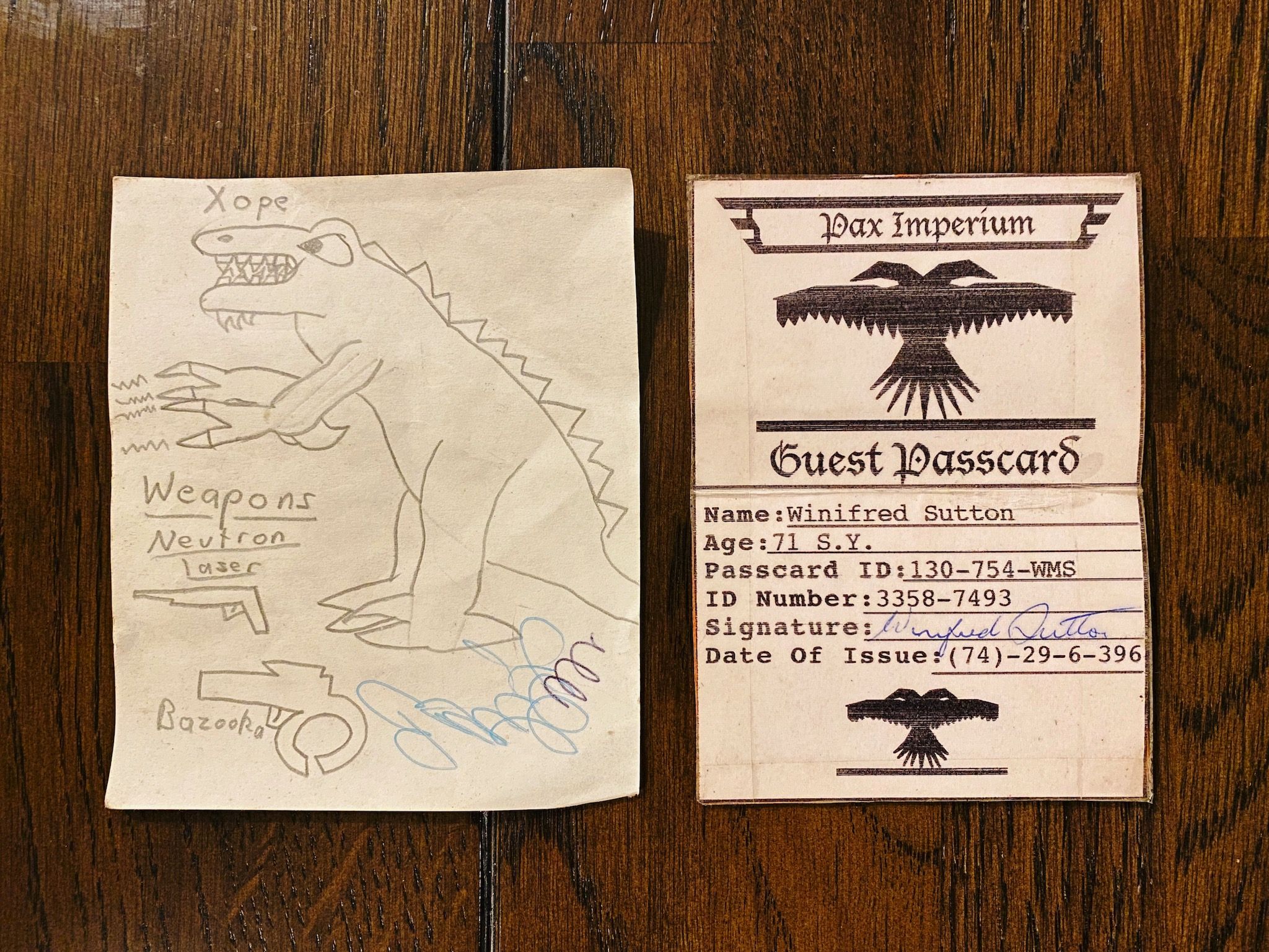 A photo showing two small pieces of paper, the first with a t-rex-type dinosaur on it with lightning coming from its front claws. Its name is "Xope" and it has two weapons, a neutron laser and a bazooka. The second paper is laminated and folds in two and says "Pax Imperium" at the top in a gothic font, with a rip-off of the 40K universe's Imperial double-headed eagle logo, with my grandma's name and age, an "ID Number", her signature, and a "Date of Issue" field that says "(74)-29-6-396".