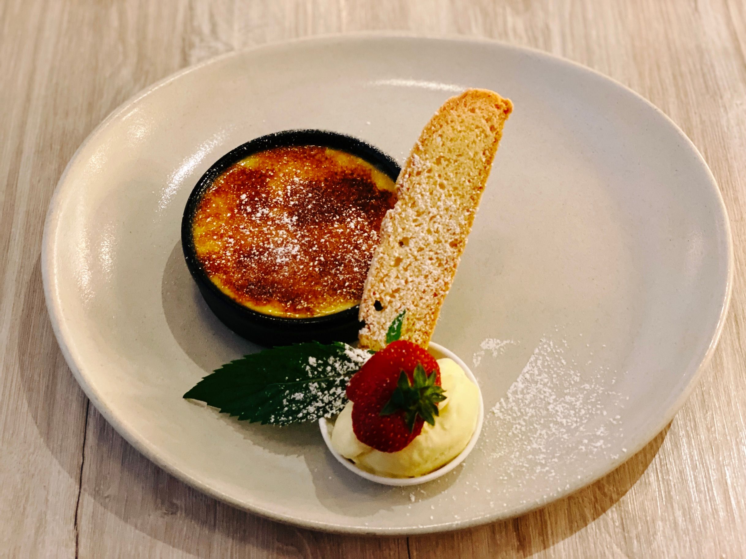 A photo of a crème brûlée alongside a scoop of vanilla bean gelato with a strawberry on top and a piece of biscotti.