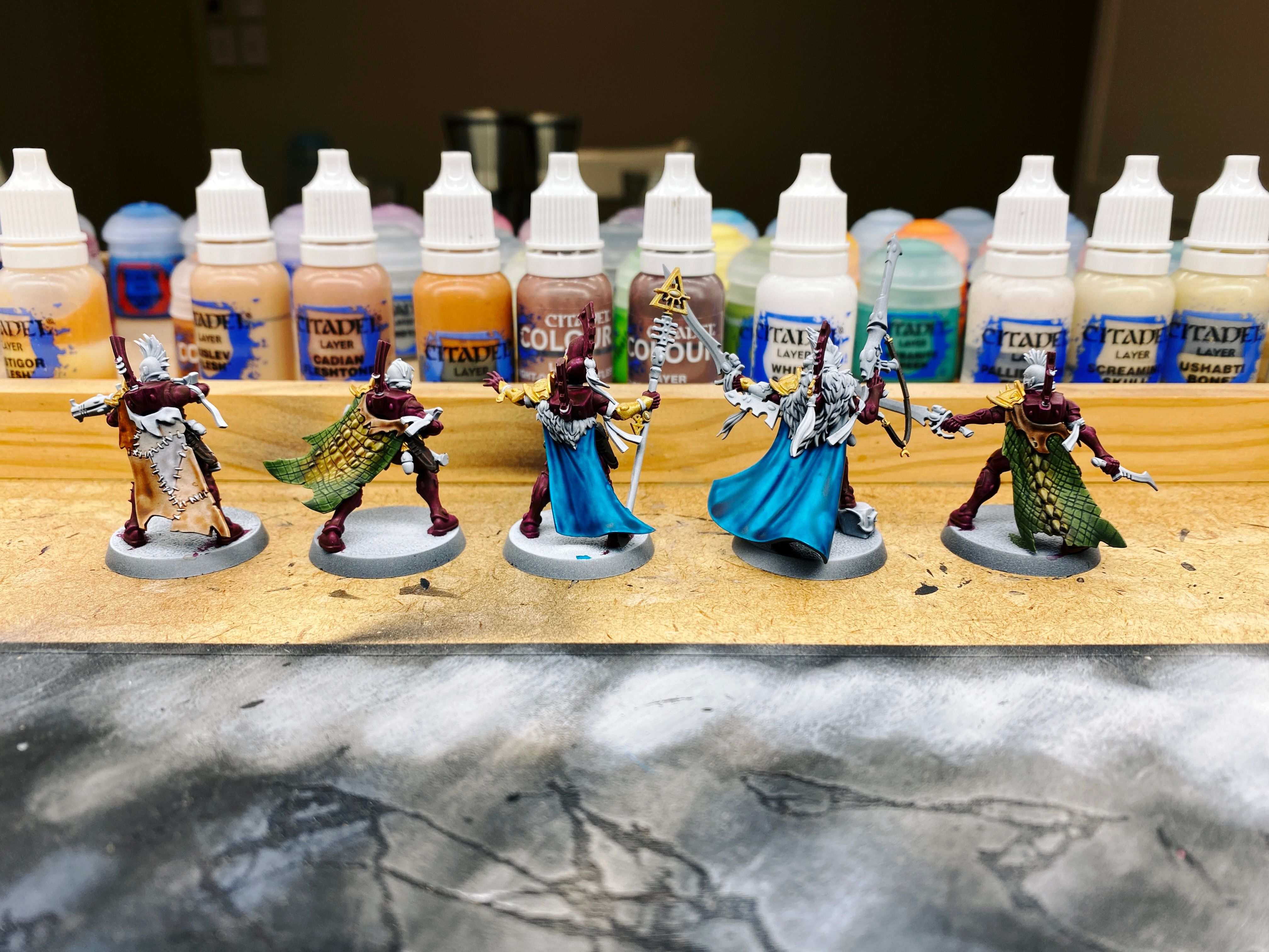 A photo of the rear of five partially-painted miniatures. They've all got long flowing cloaks, and two of them have lizard skin cloaks which are a dull green colour on the scales and faded up into a desaturated yellow colour on the knobbly bits that are obviously from the middle of the lizards' backs.
