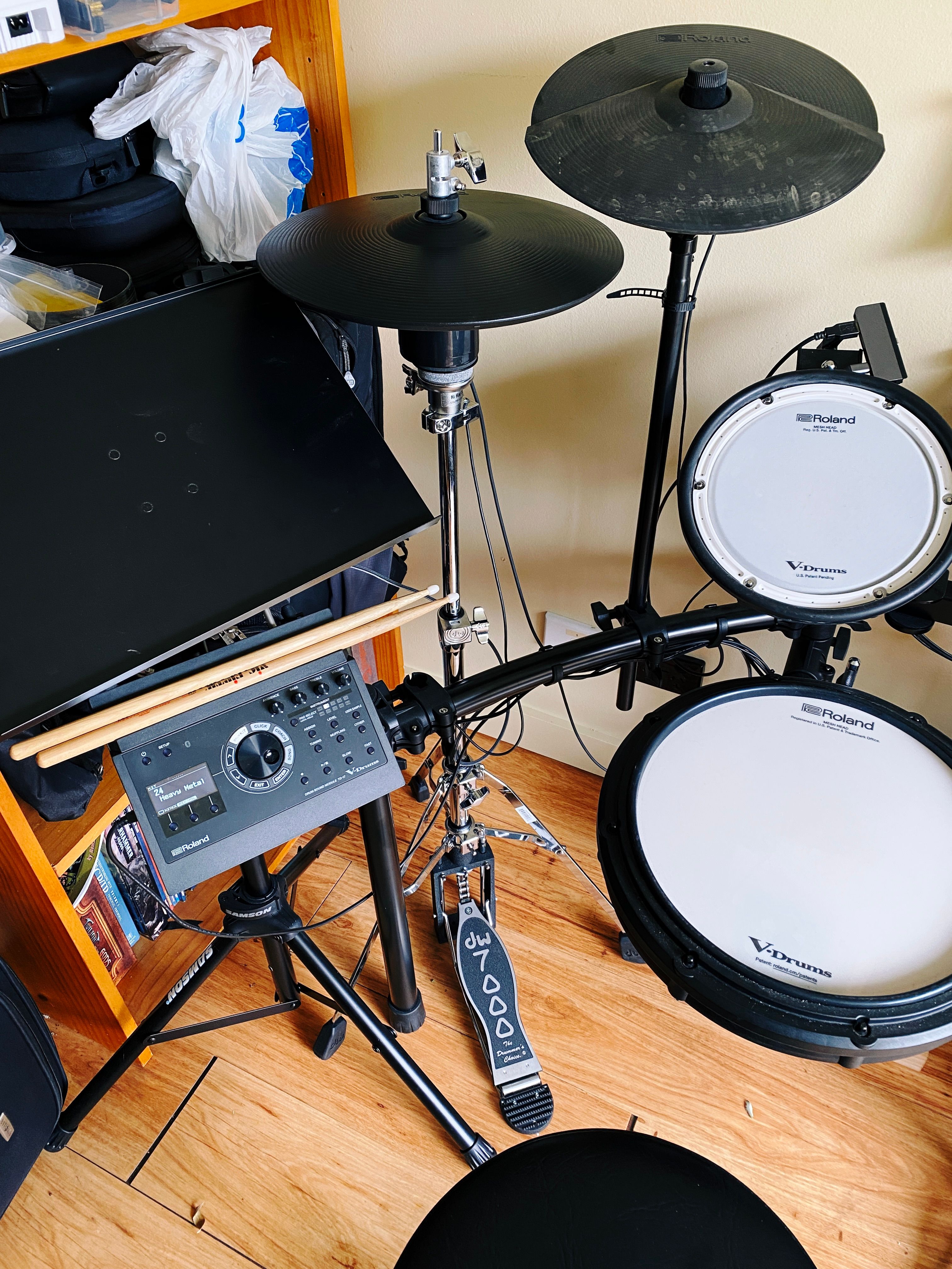 A photo of a compact electronic drum kit, with a very shiny metal acoustic hi-hat stand with a black plastic cymbal mounted on it sitting behind the kit.