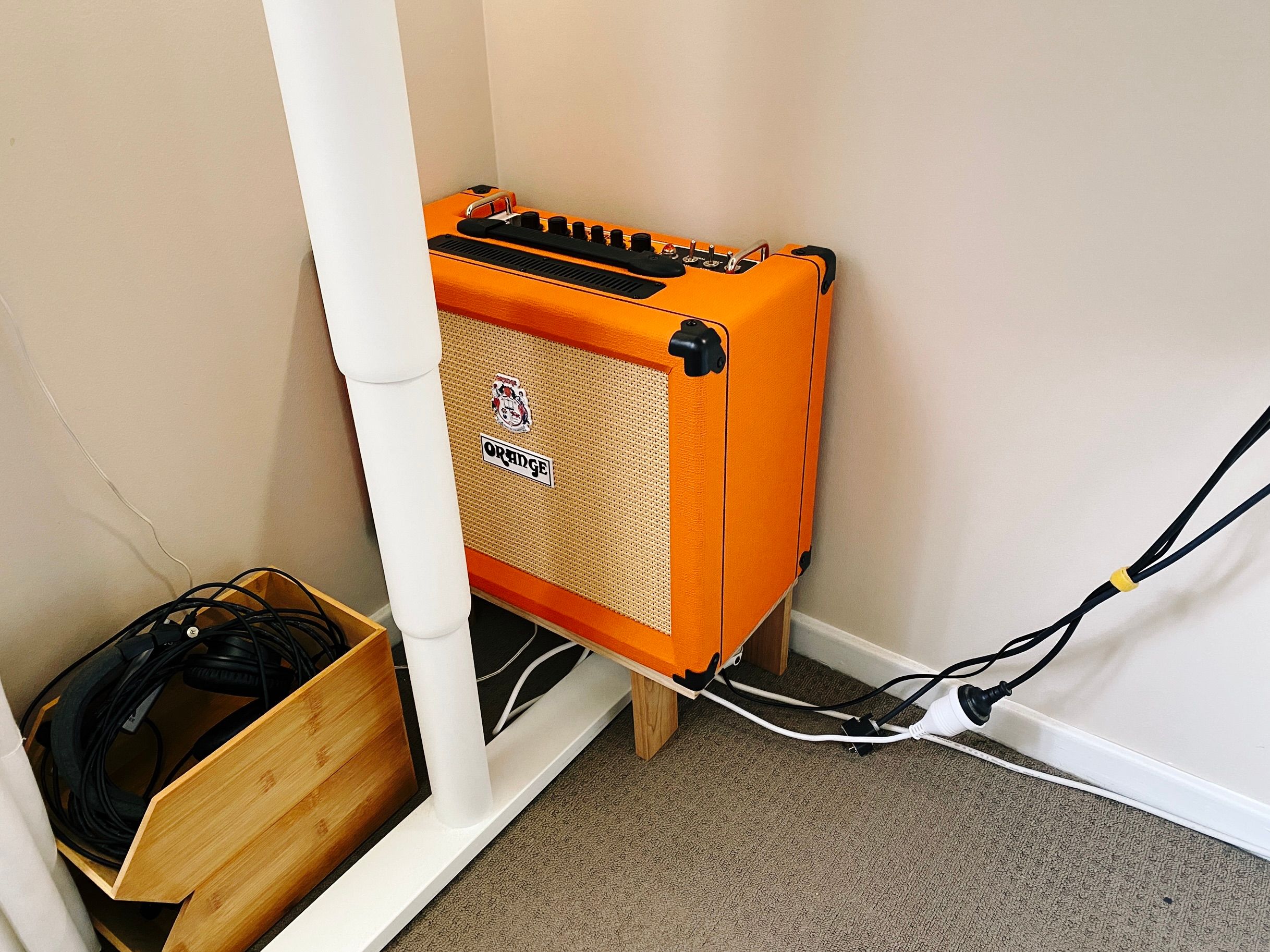 The same orange guitar amp but this time sitting on a little elevated wooden platform in the corner of a room under a standing desk.