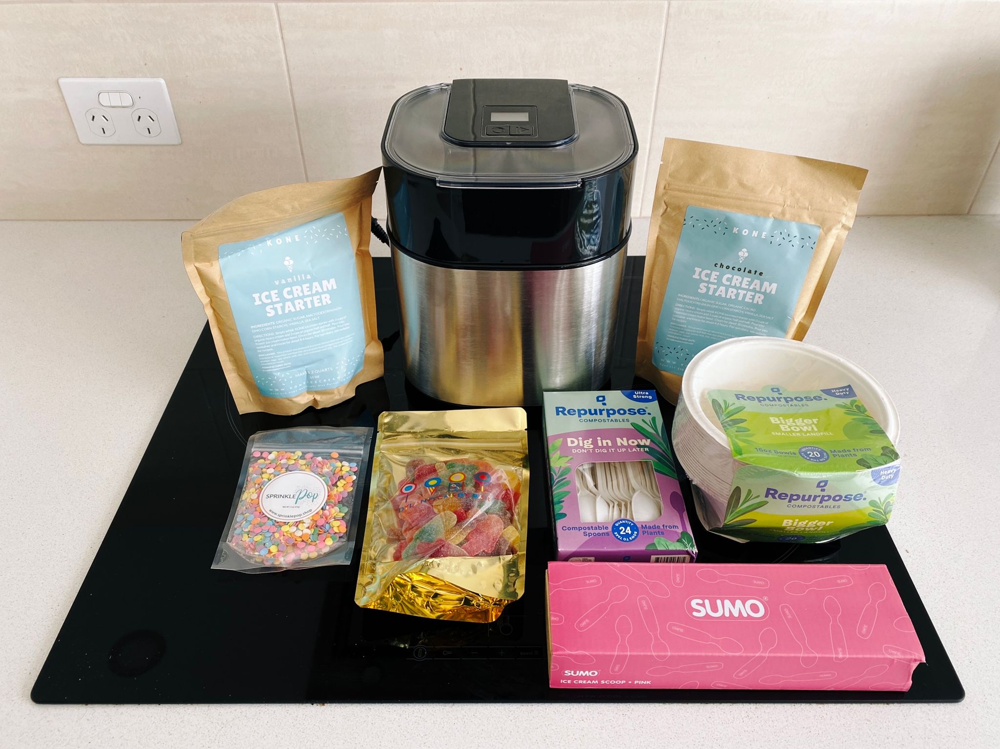 A photo of an ice cream maker along with a vanilla and chocolate ice cream starter mix, some sprinkles, a packet of lollies, an ice cream scoop, and some biodegradable bowls and spoons.