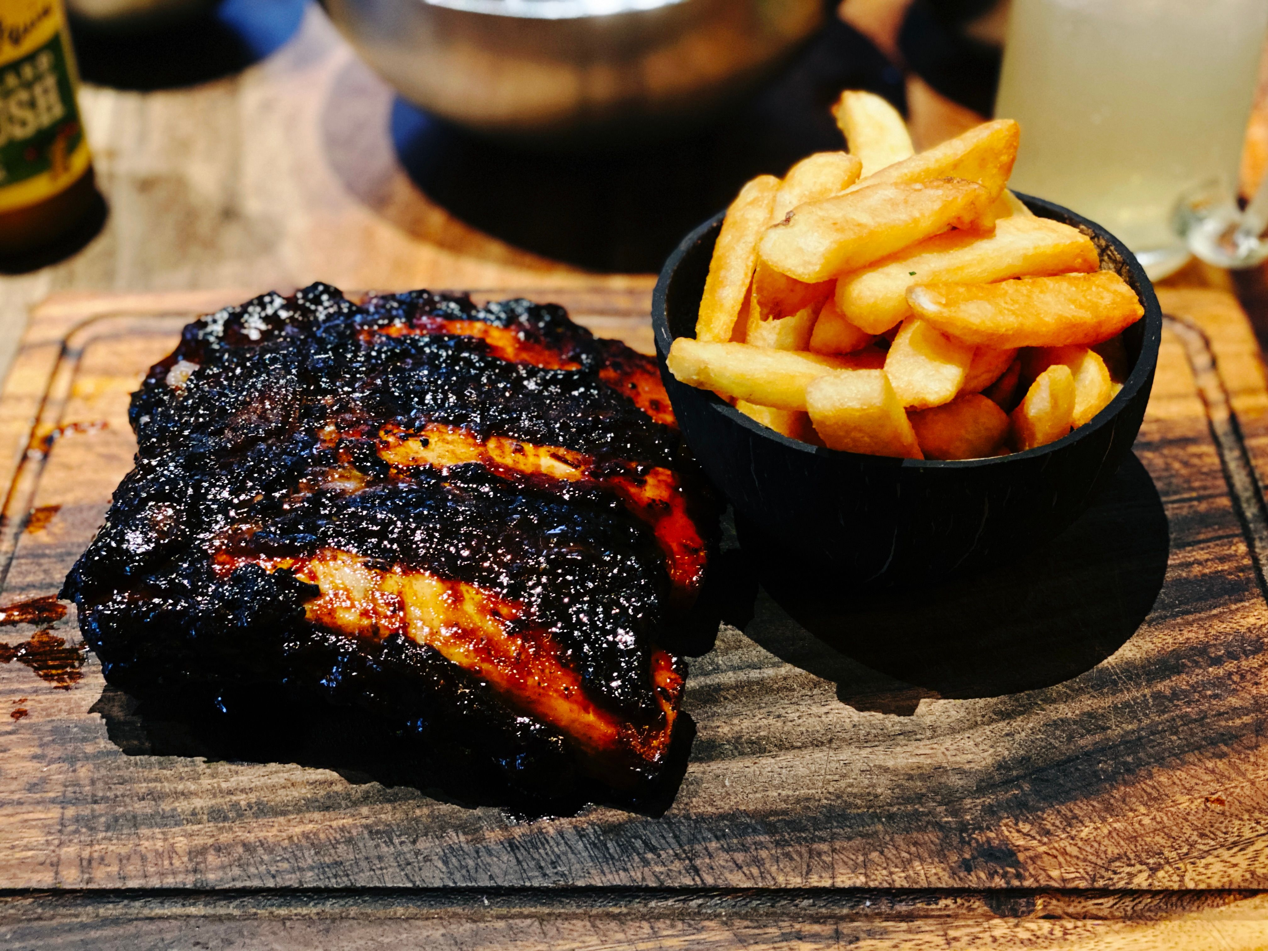 A half rack of deliciously-blackened beef ribs sitting on a wooden serving board, along with a small wooden bowl of chips.