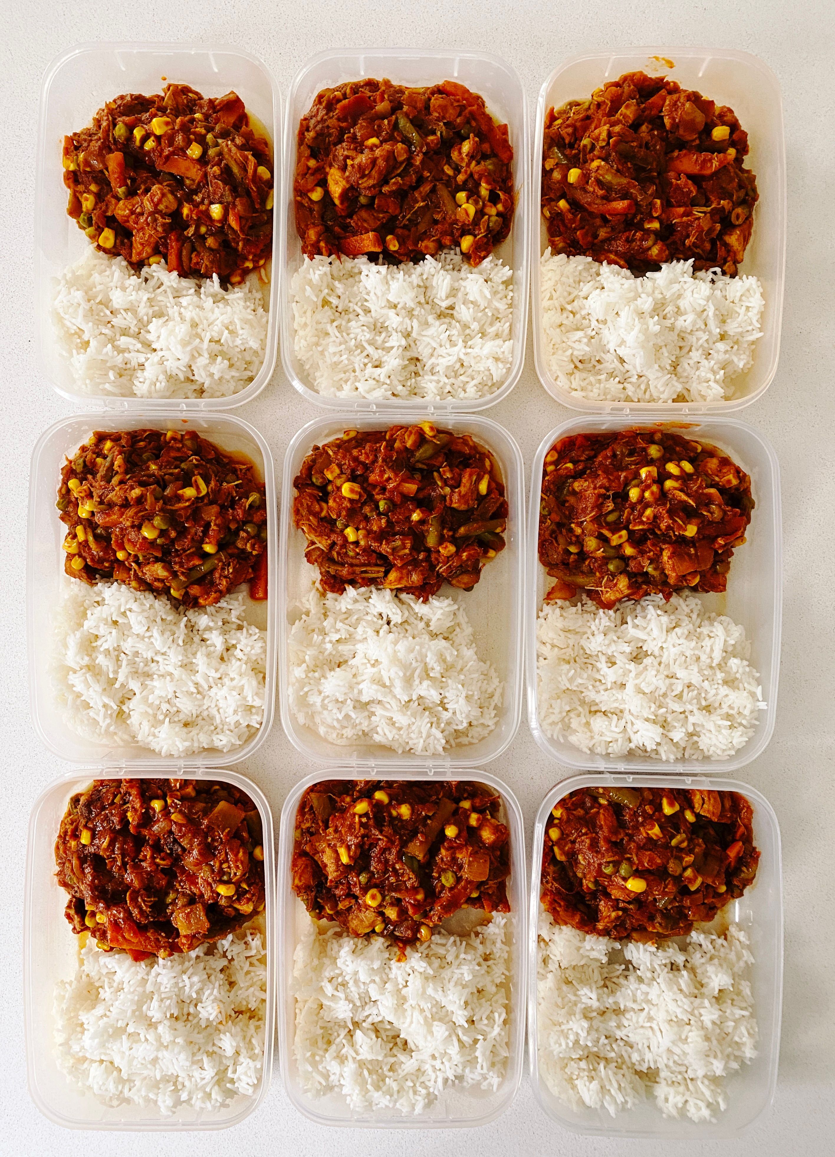 A photo of nine Tupperware containers in three-by-three pattern, each filled with half rice and half a rich red chicken curry with peas, green beans, carrots, and corn in it.