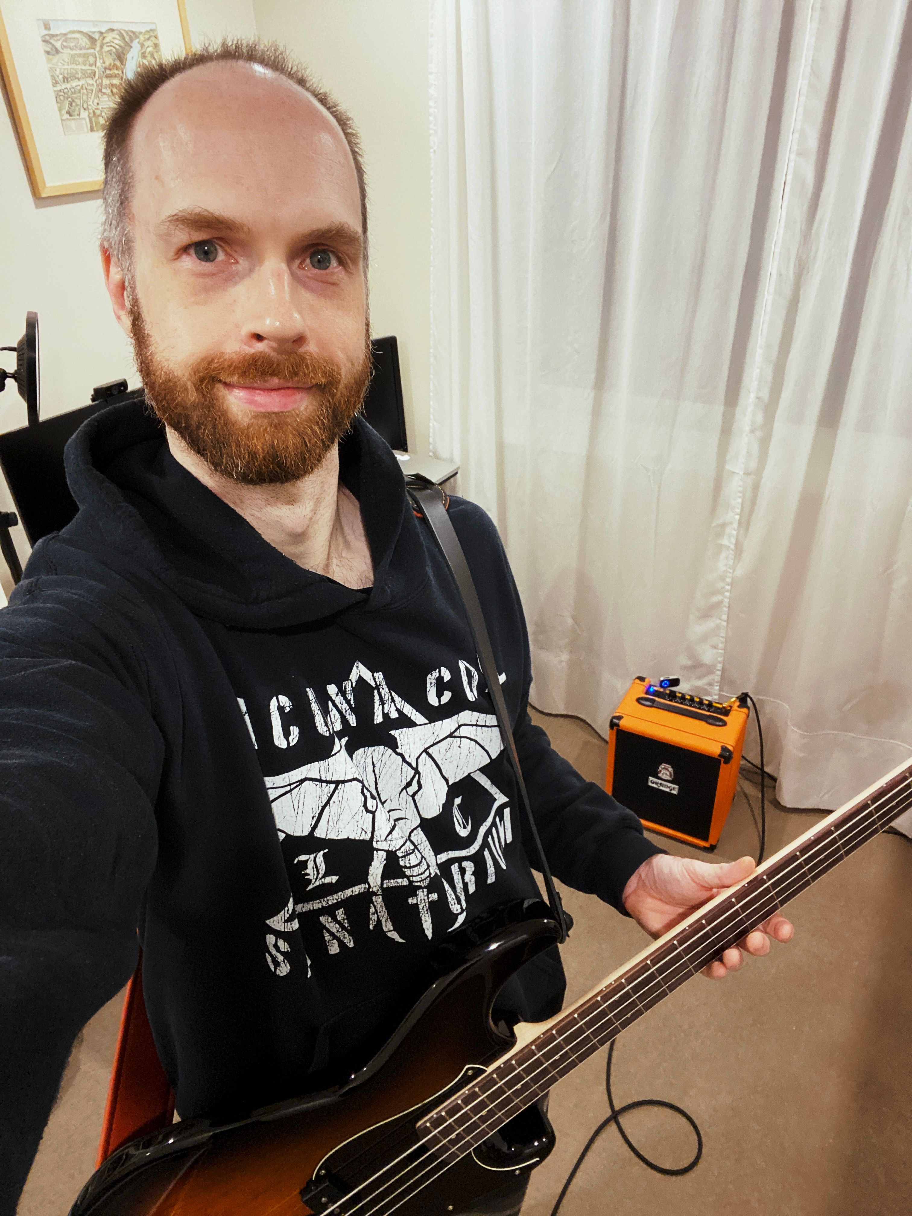 A photo of me, a white man with a beard and short hair who is in increasing need of a haircut, holding a bass guitar and smiling at the camera.
