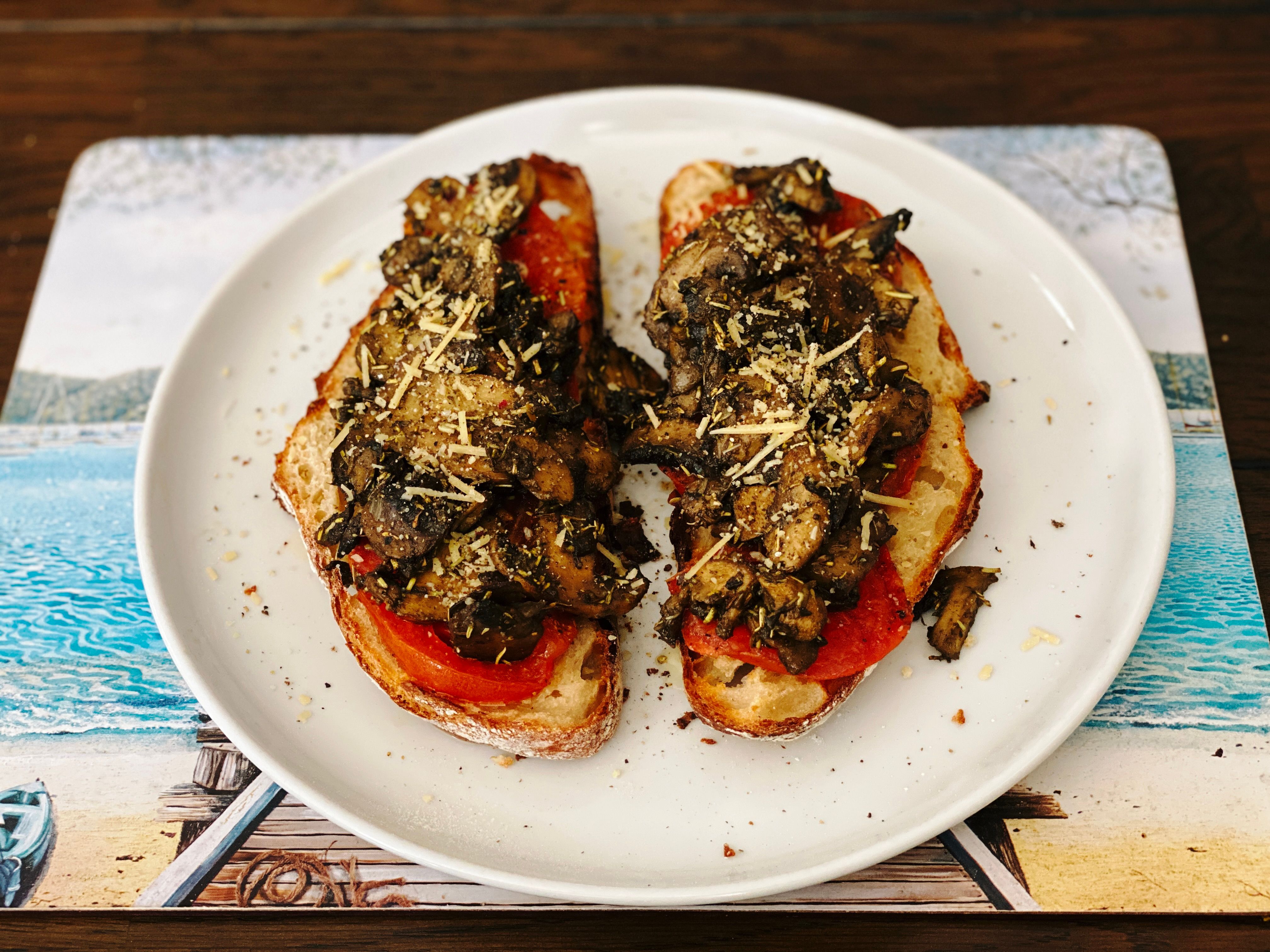 A photo of two slices of the aforementioned bread with cooked sliced tomato and mushrooms on top, sitting on a white dinner plate. There's just a little scattering of grated Parmesan cheese on top.