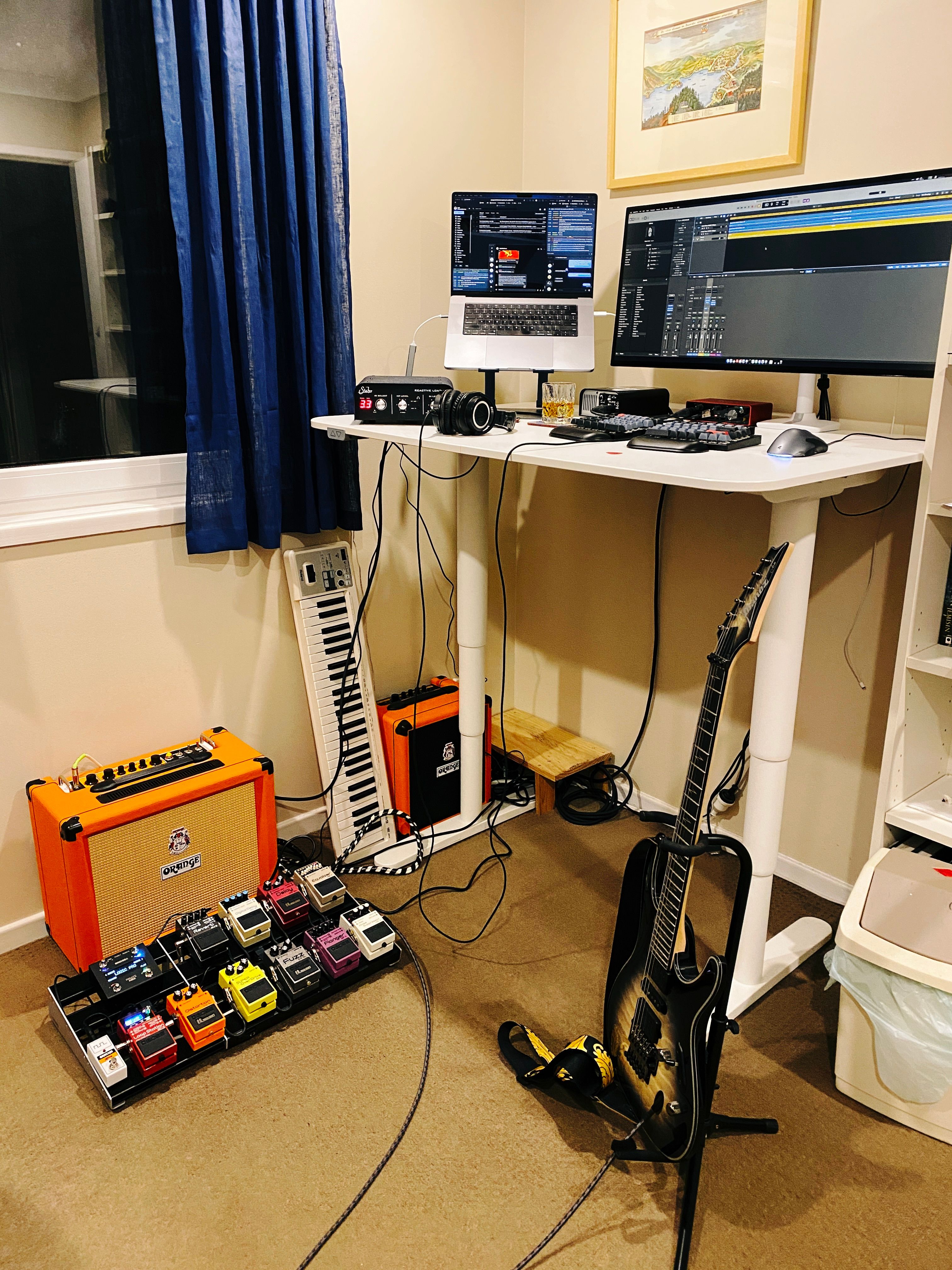 A photo of my standing desk in its upright position, with an Orange guitar amp and a pedal board with a number of guitar pedals on it sitting at the left and a guitar sitting in a guitar stand on the right. My computer has Logic Pro up on the main display.