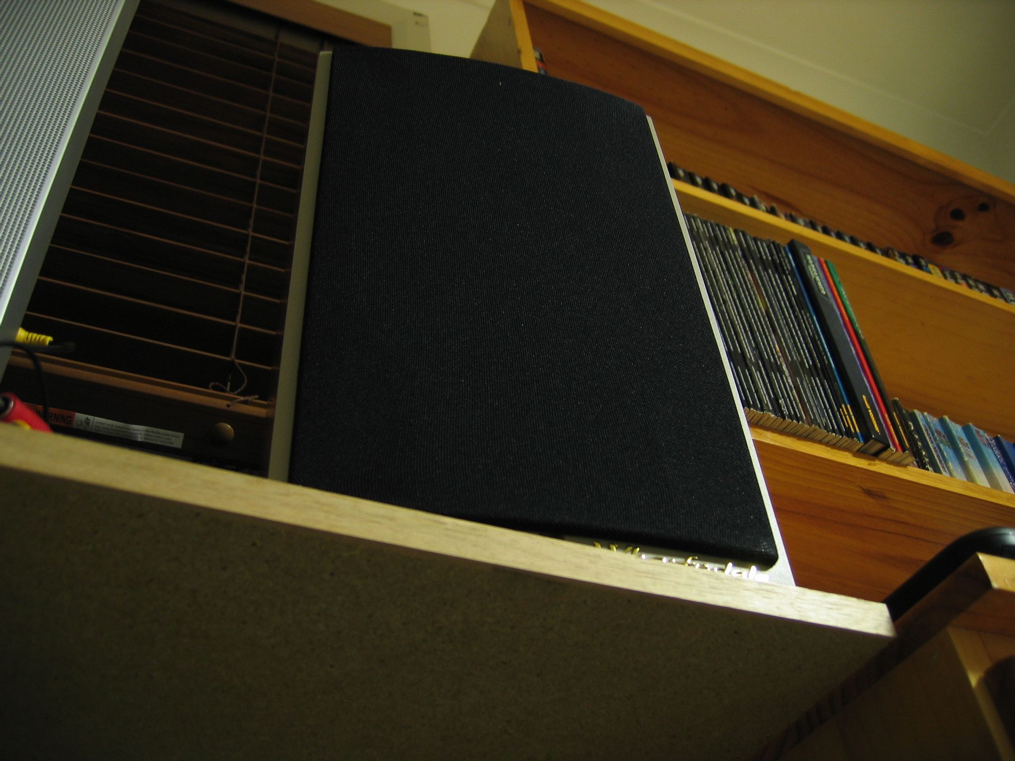 A photo taken looking up at a bookshelf speaker sitting on a shelf, and above and beyond the speaker there's a bookshelf visible with books on it.