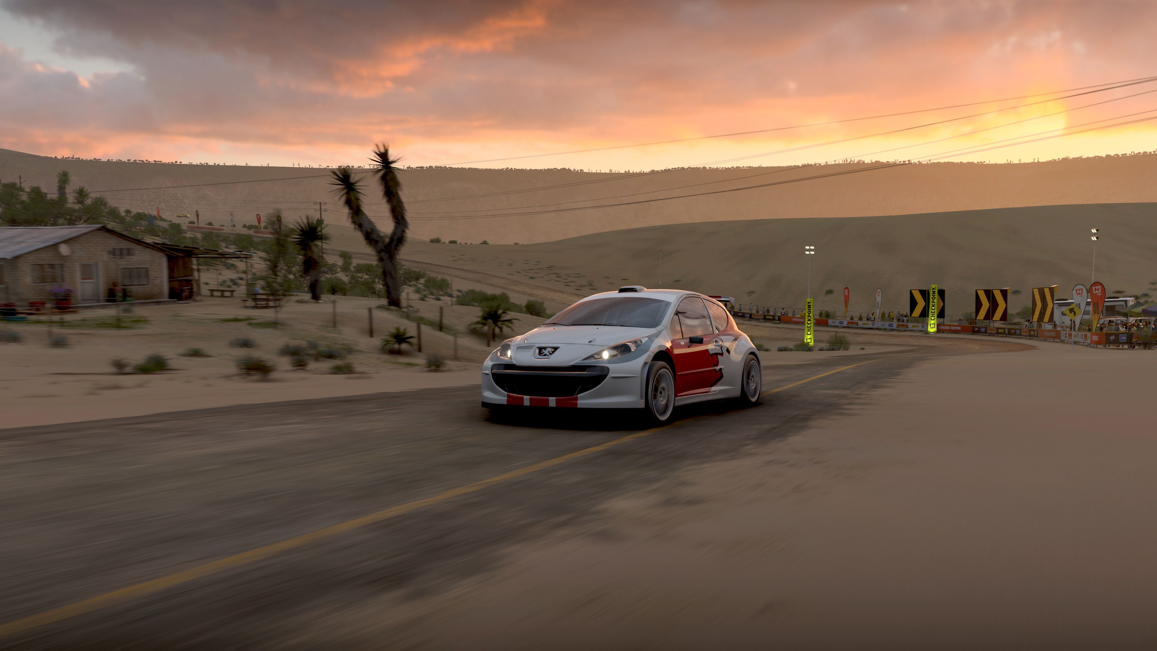 A screenshot from Forza Horizon 5, it's a front three-quarter profile shot of a white and red rally hatchback going up a very crummy-looking road in the middle of the deserts of Mexico. It's sunrise and the clouds in the distance are golden, and the photo is taken with some motion blur so it looks like the car is flying along.