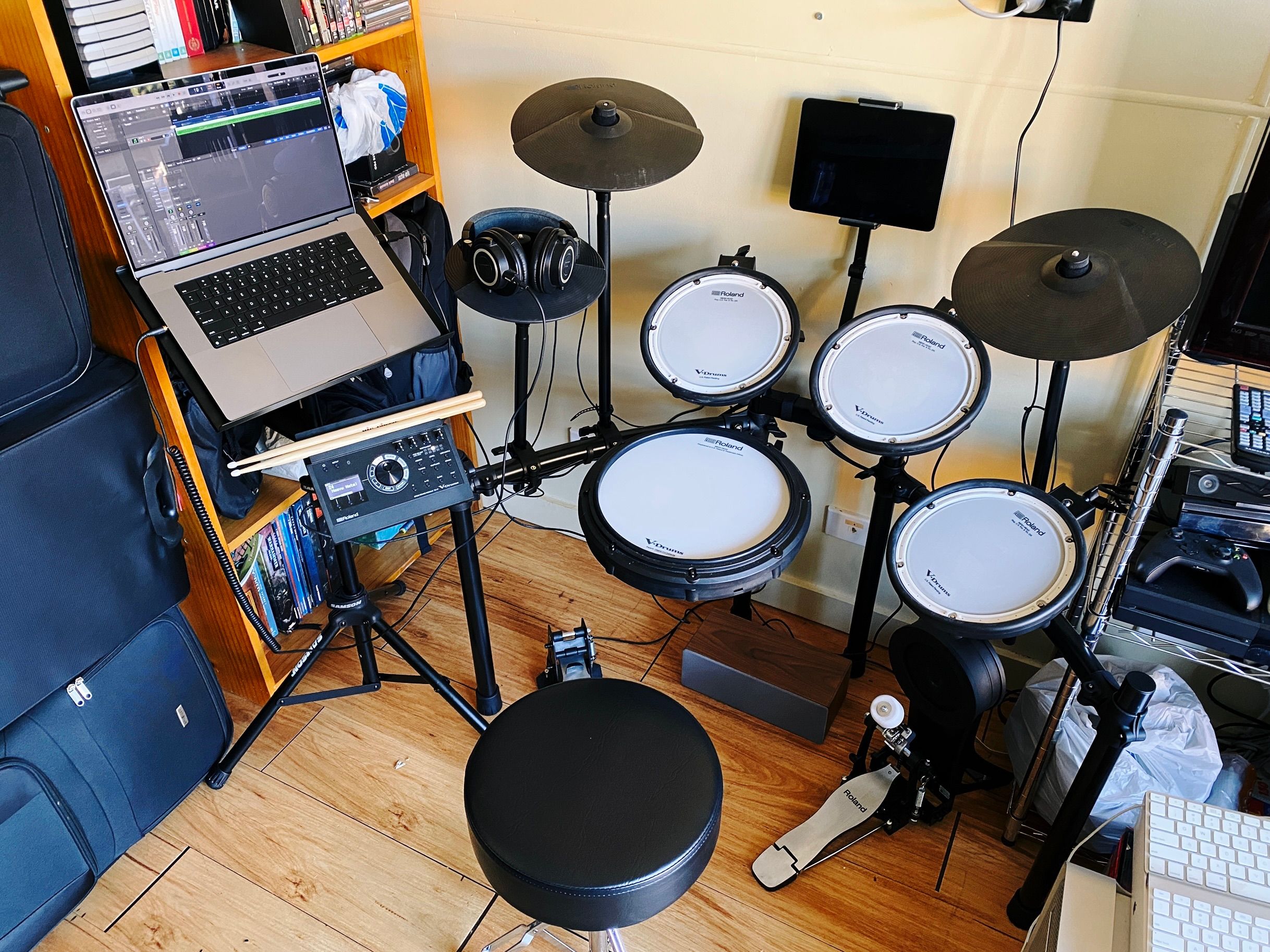 A photo of an electronic drum kit set up against a wall, with a tripod stand sitting to the left behind where the drum module is and a laptop sitting on the stand. The stand very much resembles a stand you'd put sheet music on. Immediately to the left of the drum kit and stand is a bookshelf with all sorts of non-book stuff on it, and right next to THAT is a set of luggage that'd been where I had the laptop sitting before the stand arrived.