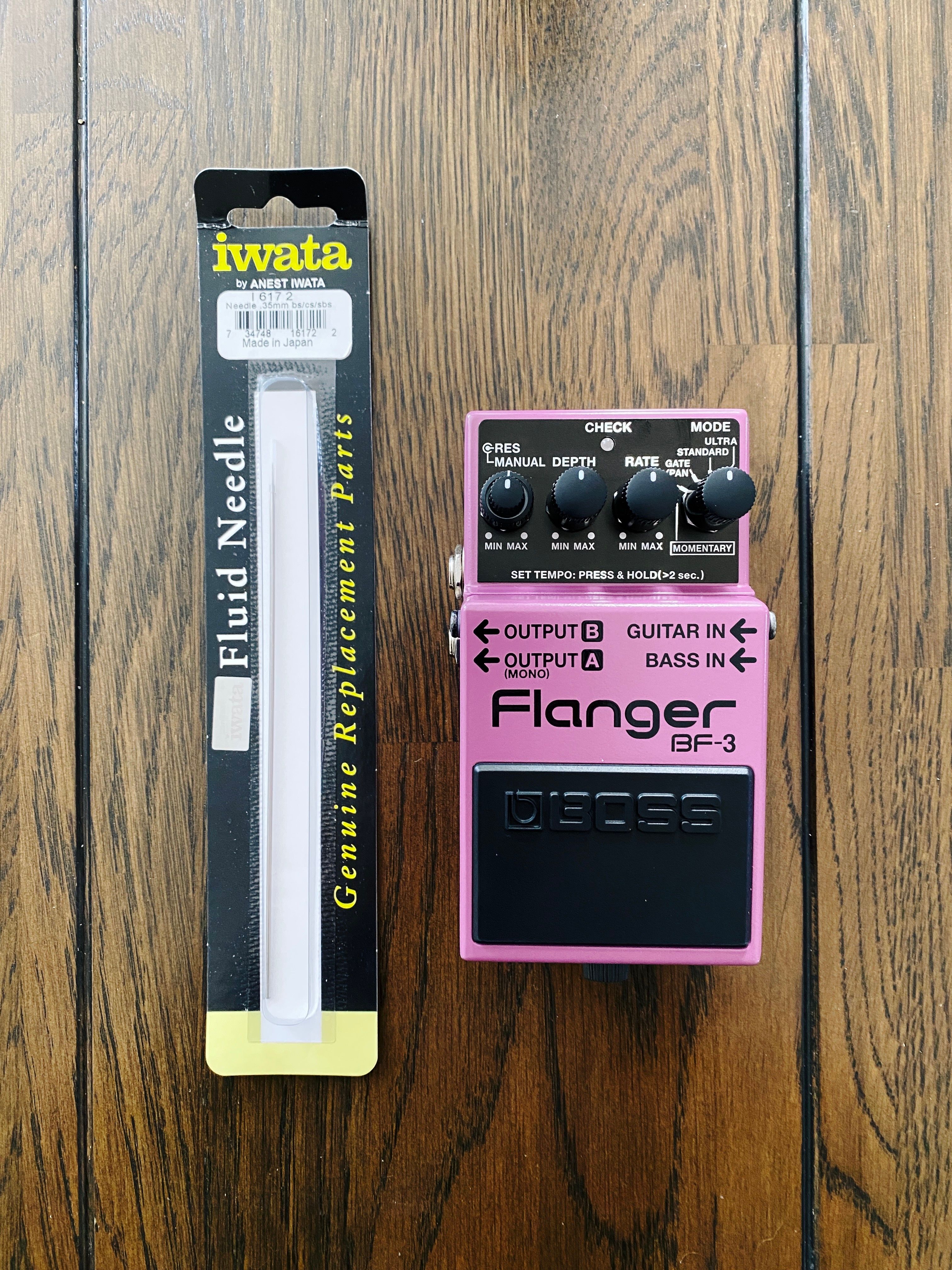 A photo of an Iwata-brand airbrush needle (literally it's just a big long metal needle with a VERY sharp tip), and a purple Boss BF-3 flanger guitar pedal, which has a stompy thing on the front to turn it on and off, and four black dials along the top.