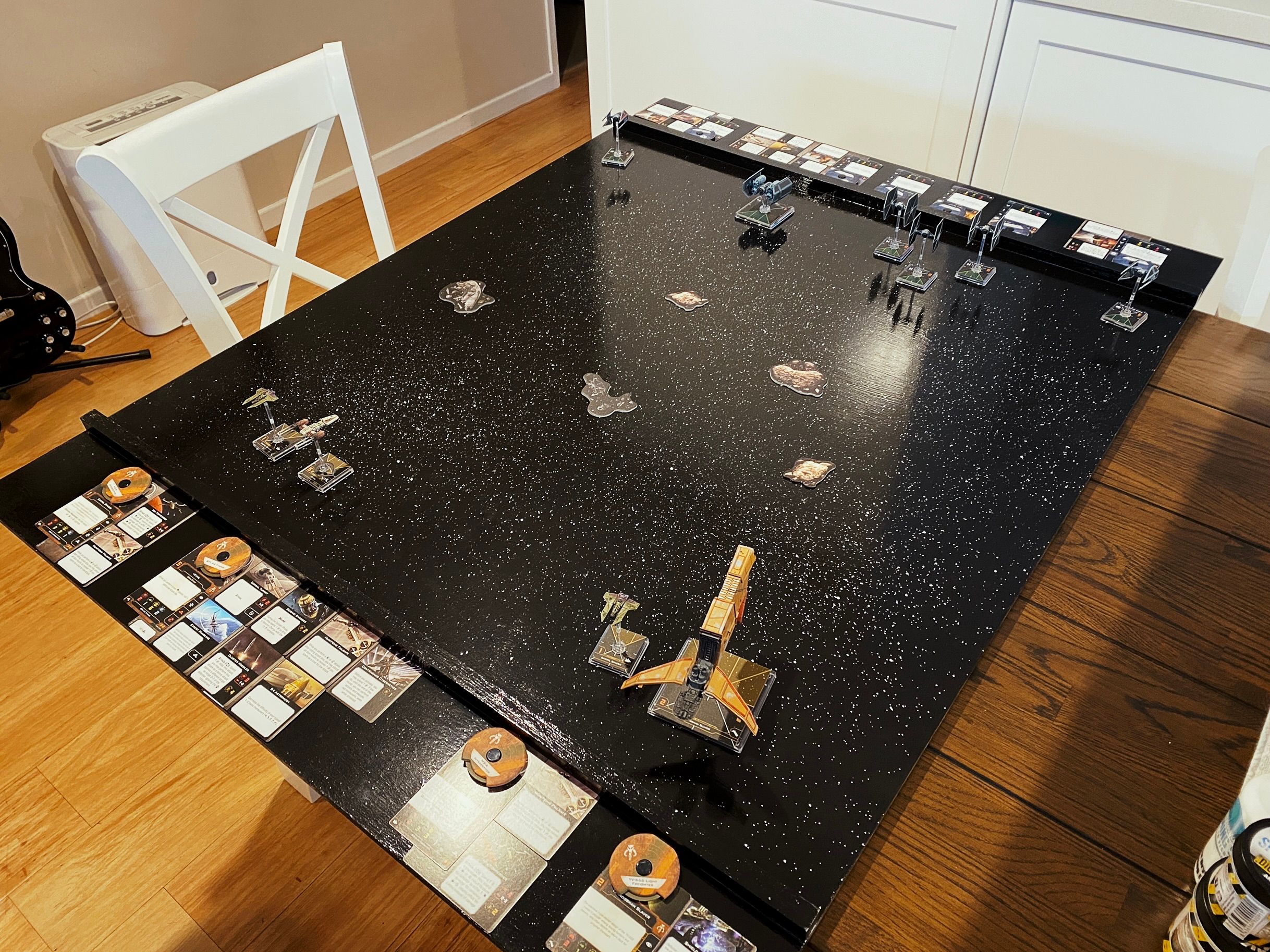 A photo of a big wooden board, about one metre by 1.3 or so, painted shiny black with a bunch of white paint spattered on it so it looks like stars. The board has miniatures from the Star Wars tabletop game "X-wing", the Empire on one side and a rag-tag squad from the Scum & Villainy faction on the other, with all their cards laid out on the section at either end that's separated from the main play area by a small raised piece of wood.