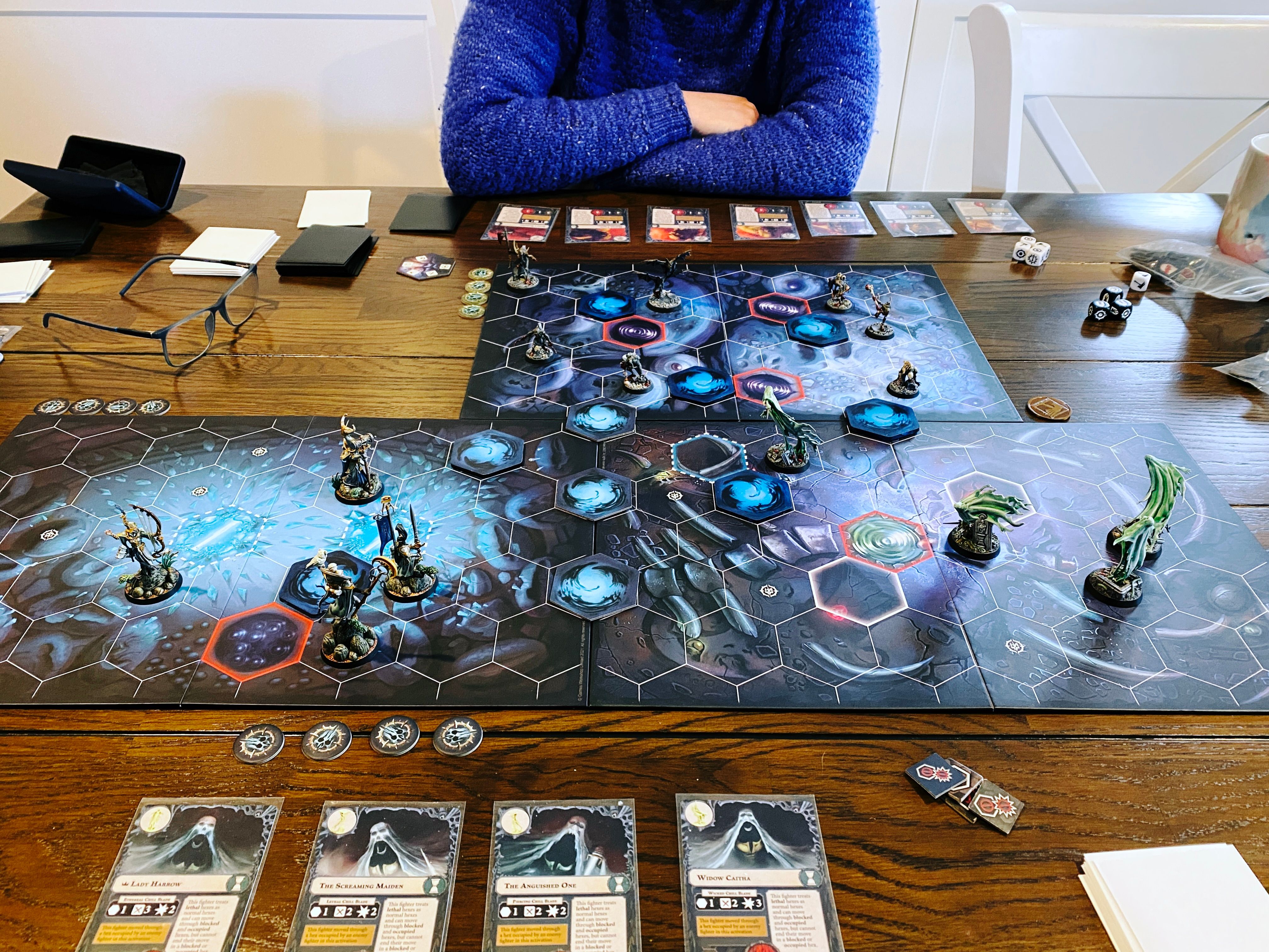 A photo of a three-player game of Warhammer Underworlds. It's a board game played on boards with hexagonal tiles, at the right are four flowing spirits in a sickly green colour, at the back are seven ghouls with rotten green-looking skin and fresh blood like they've been feeding, and at the right at four lithe and elegant elves. There's also cards laid out in front of each player, and various dice and tokens scattered about.