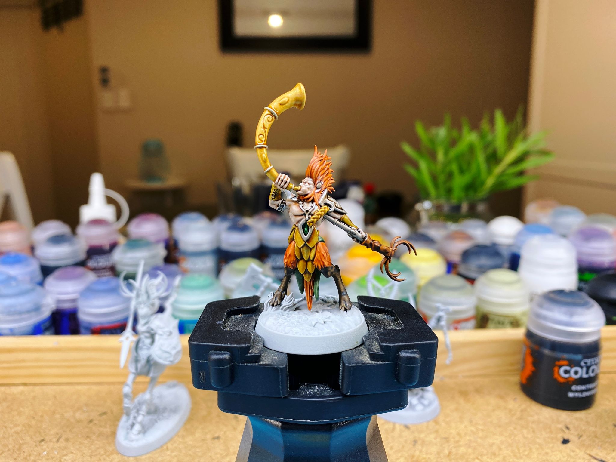 A photo of a mostly-painted miniature from Warhammer Underworlds: Beastgrave. It's a humanoid with a wild shock of orange hair, he's got backwards-jointed hoofed legs, and is blowing a very large curved golden horn.