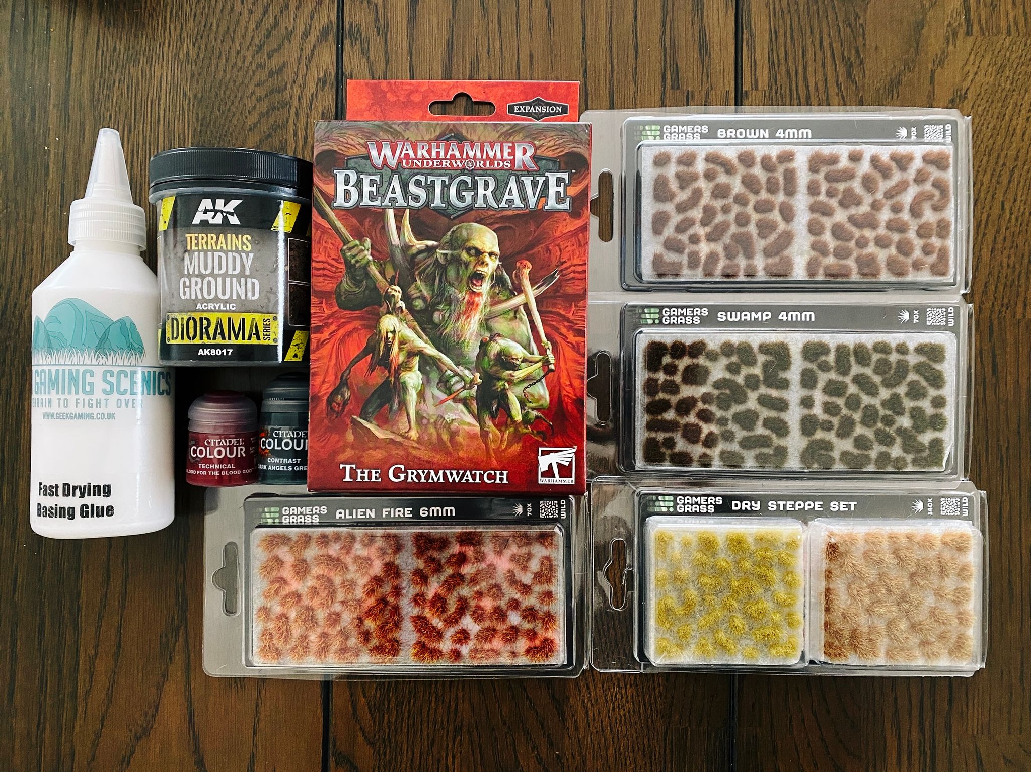 A photo of the box of the Warhammer Underworlds warband "The Grymwatch", which are essentially zombies. Also a bunch of little different coloured tufts of grass that are designed to stick onto a miniature's base and add visual interest, and a tub of "muddy ground" brown texture paint, also for basing.