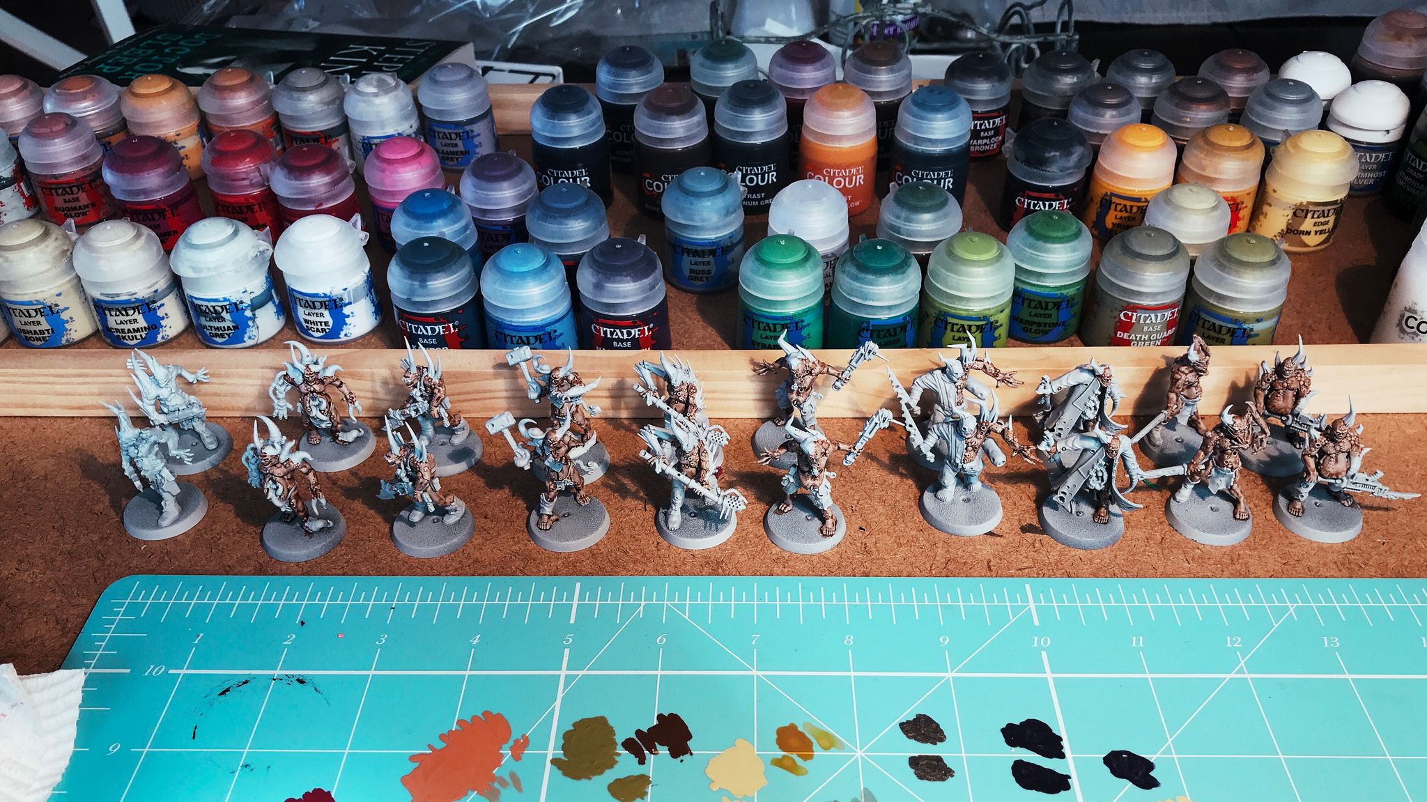Two lines of partially-painted Nurgle Poxwalkers (think zombies but with more guts hanging out of them and also lots of pustules on their skin) sitting on a desk, with a bunch of paint pots behind them.