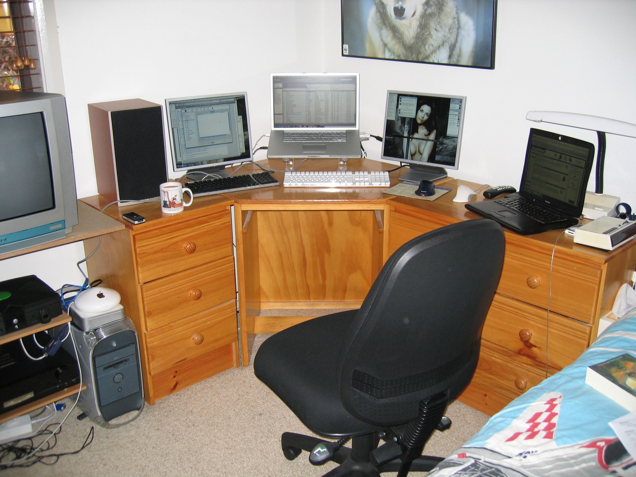 A photo of my bedroom computer setup. There's a corner desk with a 17" PowerBook on a laptop stand in the middle, flanked by 2 17" 4:3 monitors. A black Pismo-model PowerBook G3 is open to the right. On the floor to the left of the desk is a graphite Power Mac G4, with a G4 Mac mini sitting on top of it, and on top of THAT is a dome-shaped AirPort Extreme wireless router.